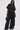 A woman poses in the HAPPY HOUR FRENCH TERRY JUMPSUIT featuring an oversized fit, wide legs, and large front pockets. She pairs it with studded black shoes, sunglasses, and a black handbag against a plain white background.