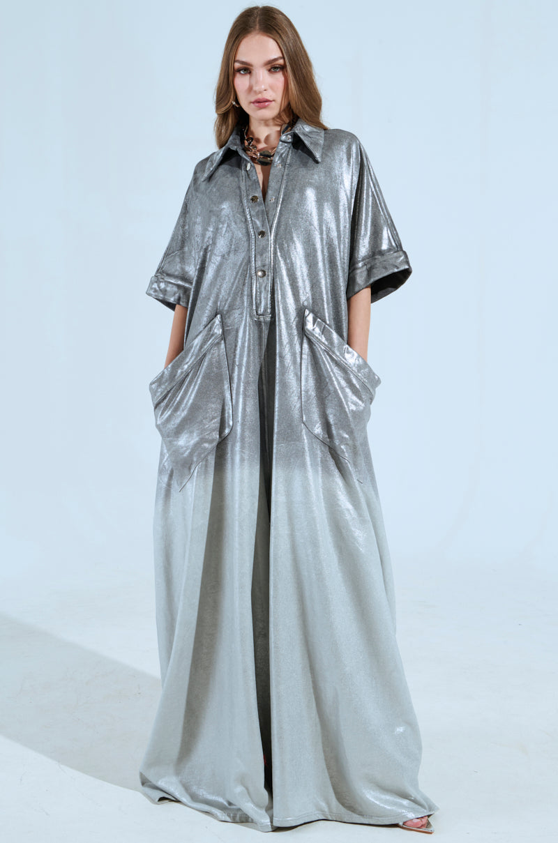 A person stands confidently in a long, silver ombre jumpsuit reminiscent of chic French Terry style, featuring short sleeves and large pockets. With hands in pockets, they pose effortlessly against a plain white background. Product: LIFE GOES ON OMBRE FRENCH TERRY JUMPSUIT.
