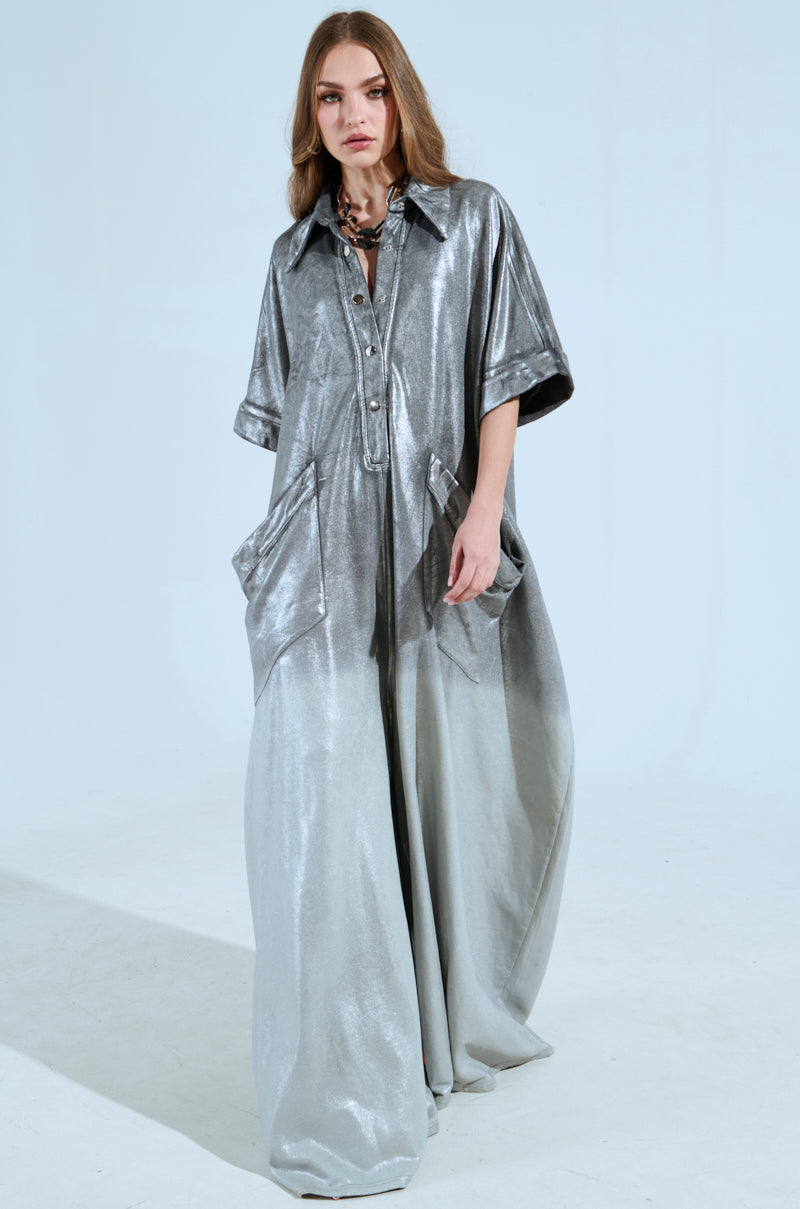 A woman poses against a plain backdrop in the LIFE GOES ON OMBRE FRENCH TERRY JUMPSUIT, featuring a flowing metallic silver design with large pockets. She has long, wavy hair and accessorizes with a chunky necklace, while the jumpsuit's short sleeves and button-up front add elegance to her chic ensemble.