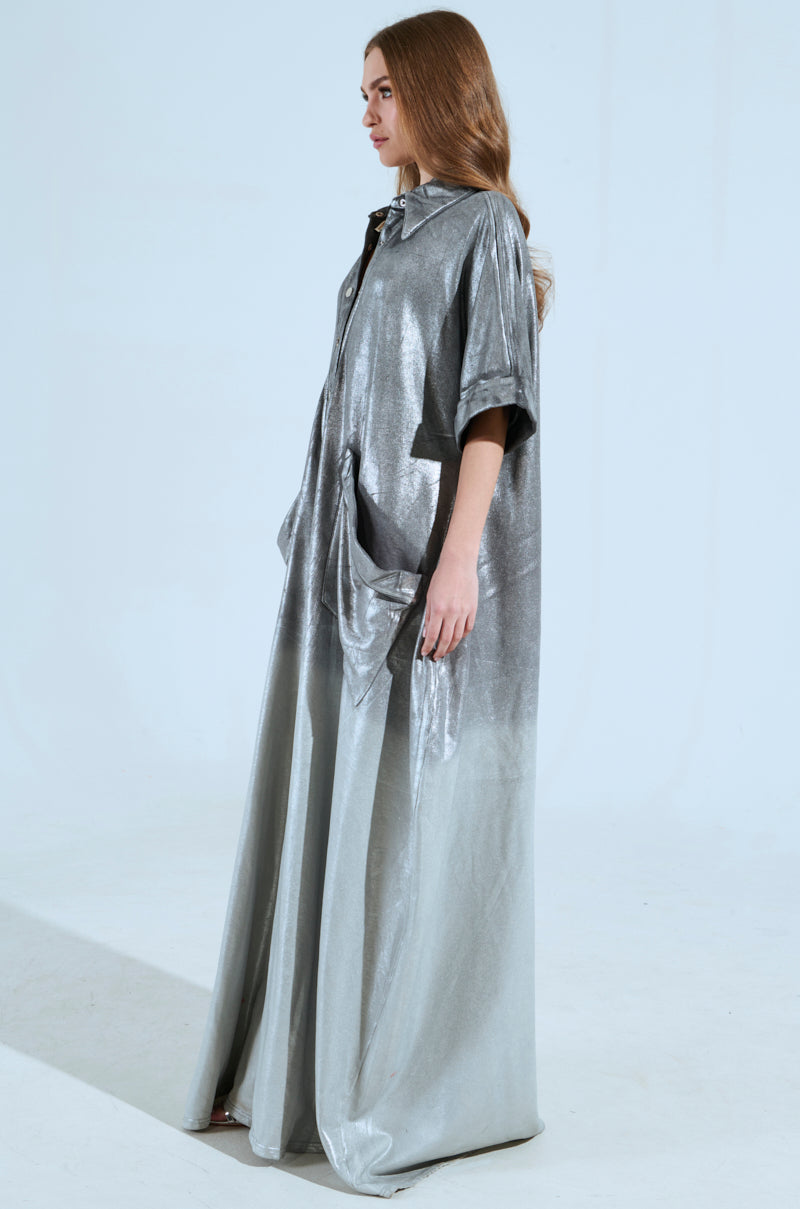 A person with long hair wears the LIFE GOES ON OMBRE FRENCH TERRY JUMPSUIT, standing in a minimalistic, neutral-toned background. The jumpsuit features short sleeves and a metallic silver design with a shiny texture, as they look to the side.