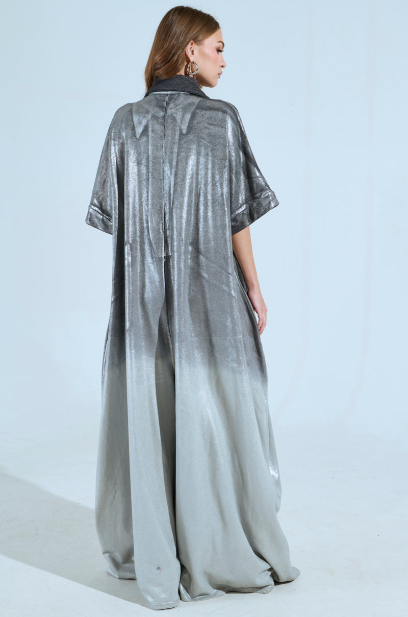 A woman with long hair showcases the drape and shine of the LIFE GOES ON OMBRE FRENCH TERRY JUMPSUIT, which transitions to a lighter shade at the hem. She stands against a light blue background, facing to the side.