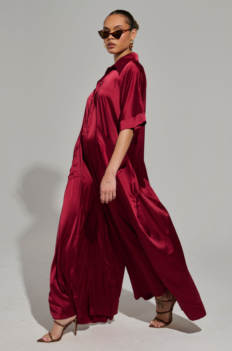Clad in the stunning HAPPY HOUR SATIN JUMPSUIT DRESS, a confident individual strides forward, the deep red fabric flowing elegantly. They accessorize with chic brown sunglasses and matching strappy heels, while their hair is neatly pulled back. The subtlety of a light gray background enhances the ensemble's elegance.