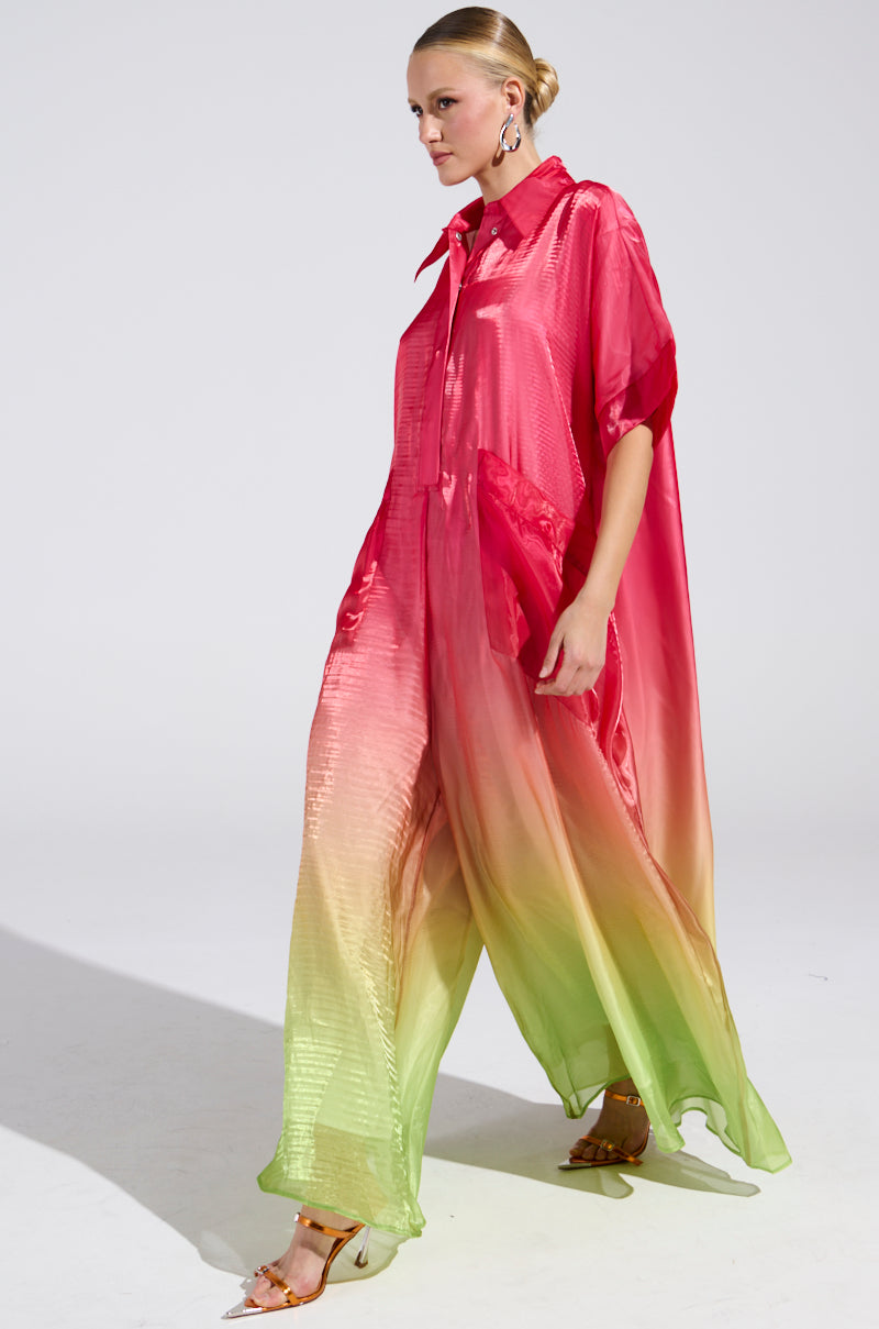 A person confidently wears the Isla Ombre Oversized Satin Jumpsuit in pink, complemented by gold heels and hoop earrings, standing elegantly against a plain white studio setting.