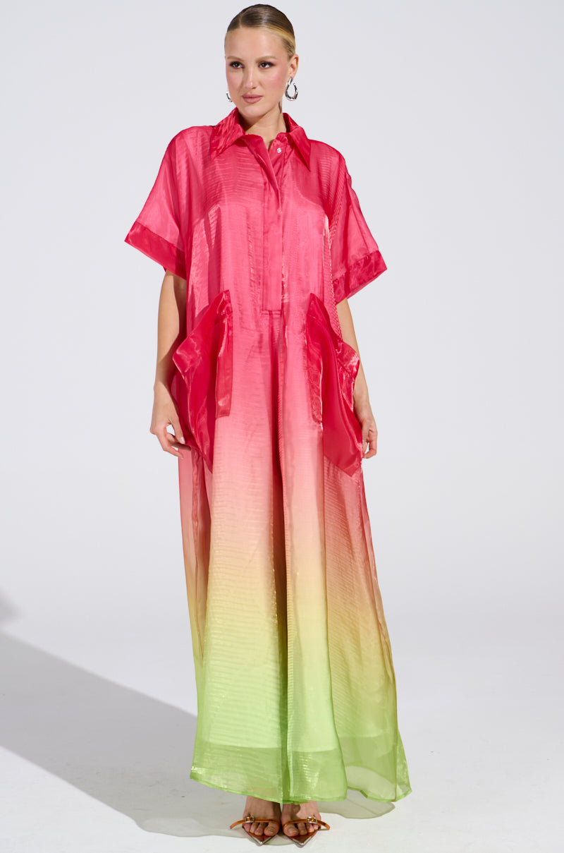 Someone stands against a plain background, wearing the ISLA Ombre Oversized Satin Jumpsuit in pink. This jumpsuit showcases a vibrant gradient design from pink to green and includes two large waist pockets. Their hair is styled up.