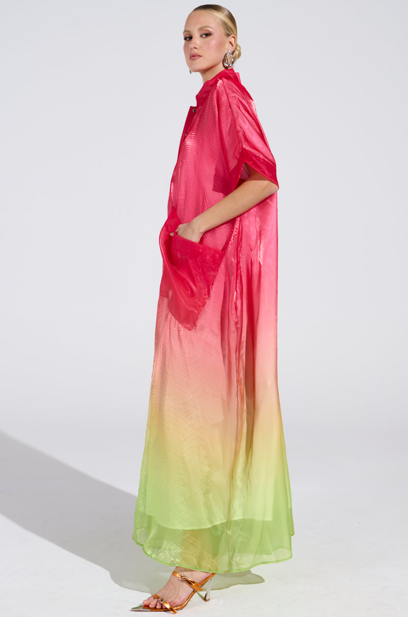 A model showcases the ISLA Ombre Oversized Satin Jumpsuit in pink, transitioning to green, with short sleeves and pockets. She pairs it with gold high-heeled sandals and sleek hair against a well-lit backdrop.