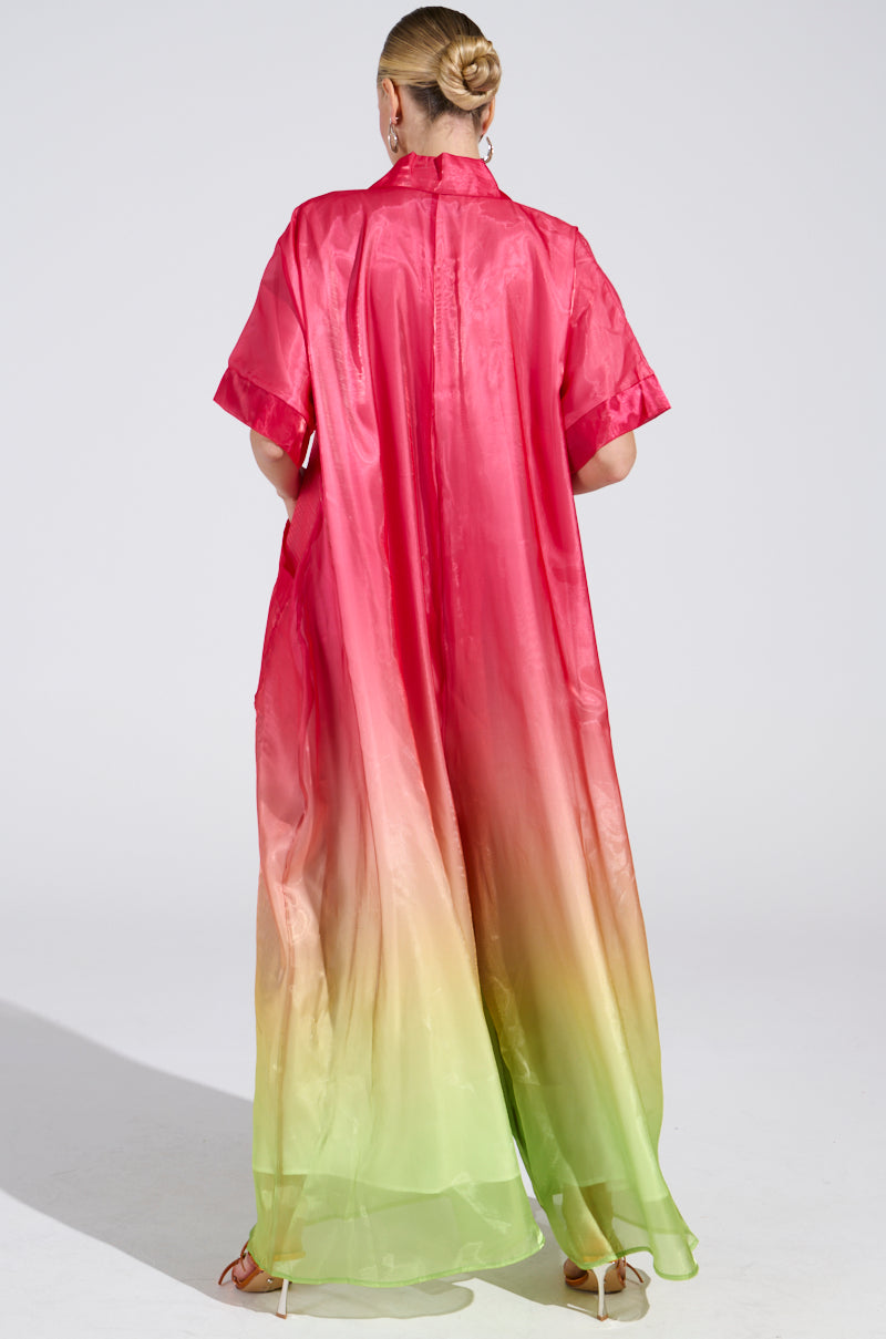 The person, with blonde hair in a bun, faces away from the camera wearing the ISLA Ombre Oversized Satin Jumpsuit in Pink with short sleeves and a pink-to-green gradient, paired with strappy sandals.