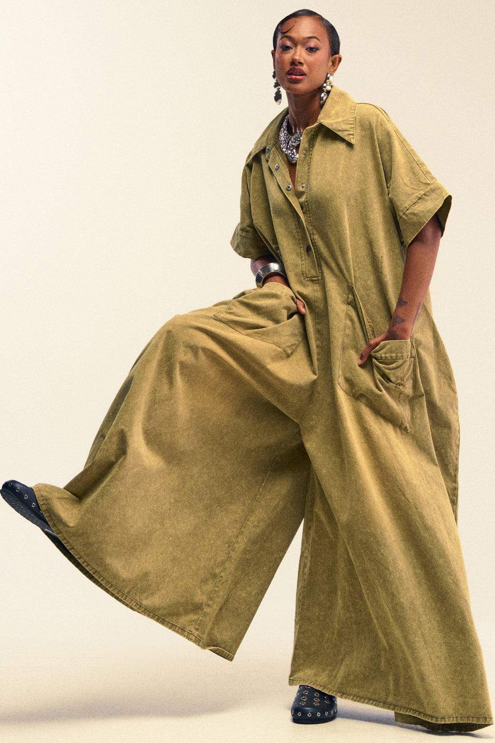 A woman poses confidently in THE BEST OVERSIZED DENIM JUMPSUIT IN OLIVE, featuring a relaxed fit with wide legs, short sleeves, and large pockets. She accessorizes with bold jewelry and black shoes against a neutral background.