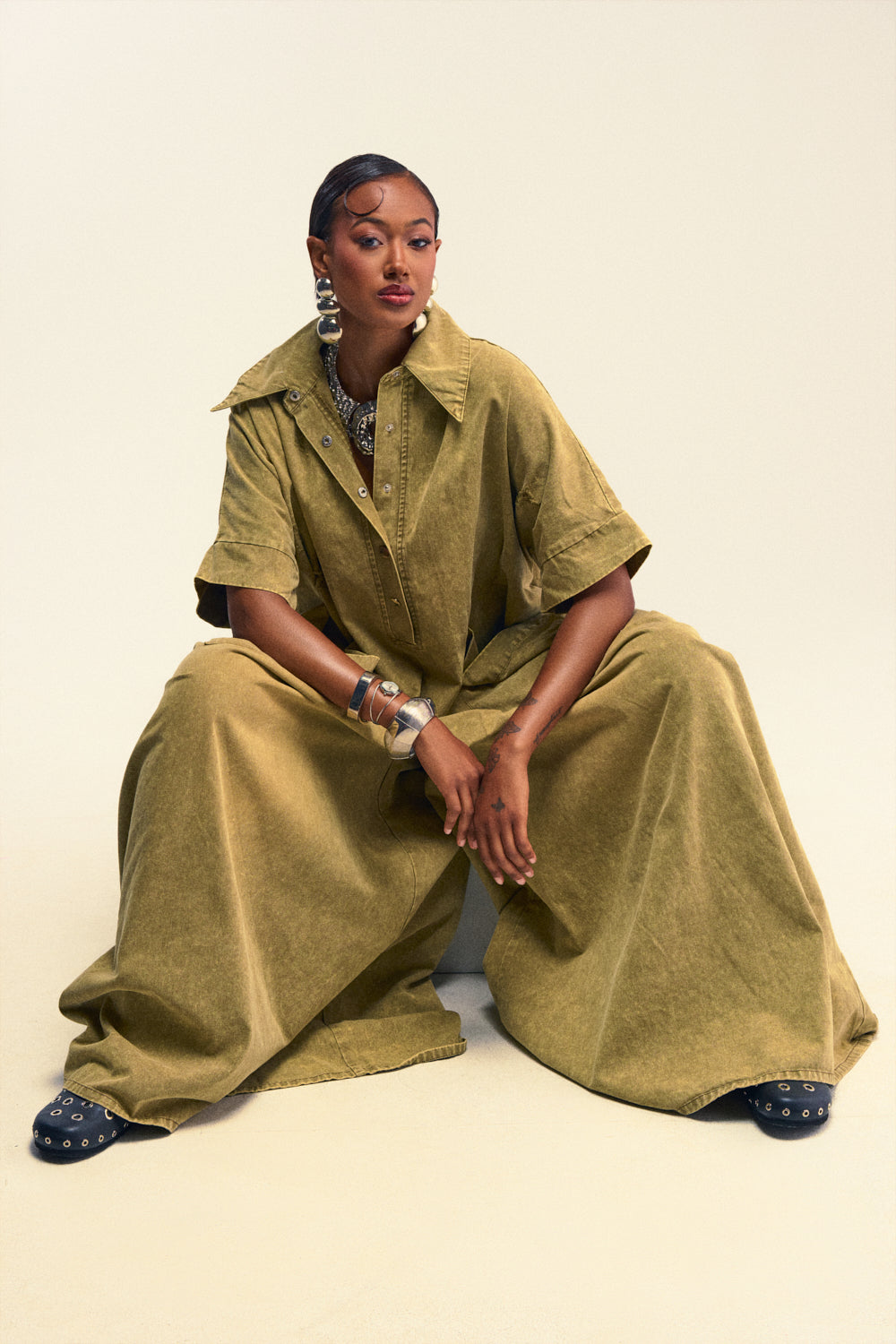 A woman sits against a plain background, confidently looking at the camera with her hands resting in front of her. She wears THE BEST OVERSIZED DENIM JUMPSUIT IN OLIVE, paired with large earrings and multiple bracelets.