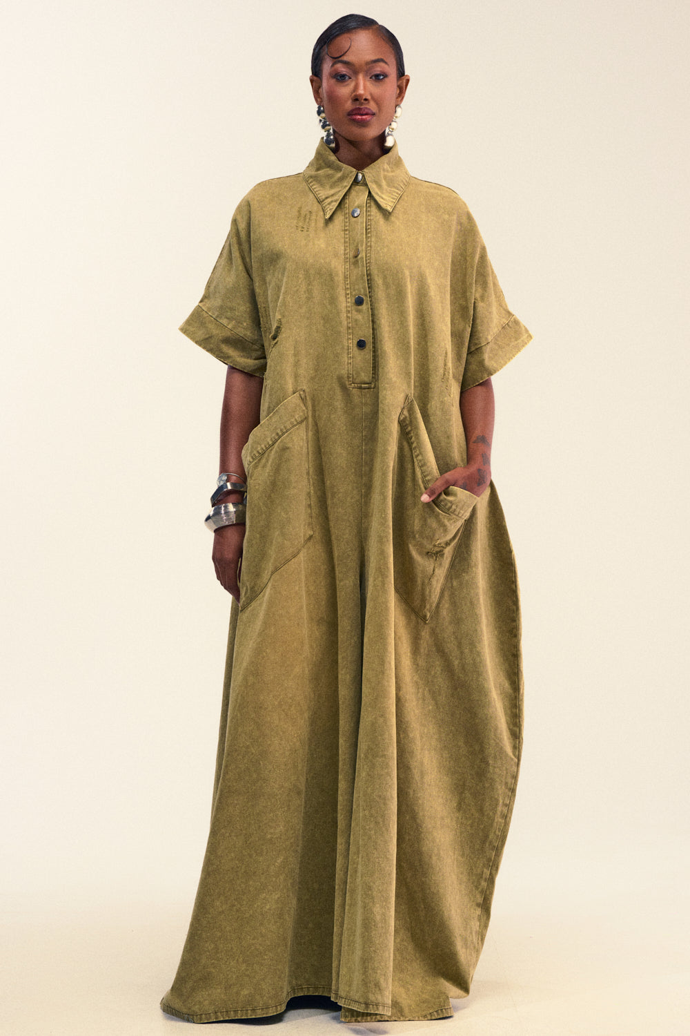Wearing THE BEST OVERSIZED DENIM JUMPSUIT IN OLIVE—with its loose, relaxed fit and practical front pockets—a person poses against a plain light background, their look styled with rolled sleeves and metallic jewelry for an effortless touch of elegance reminiscent of a maxi dress.