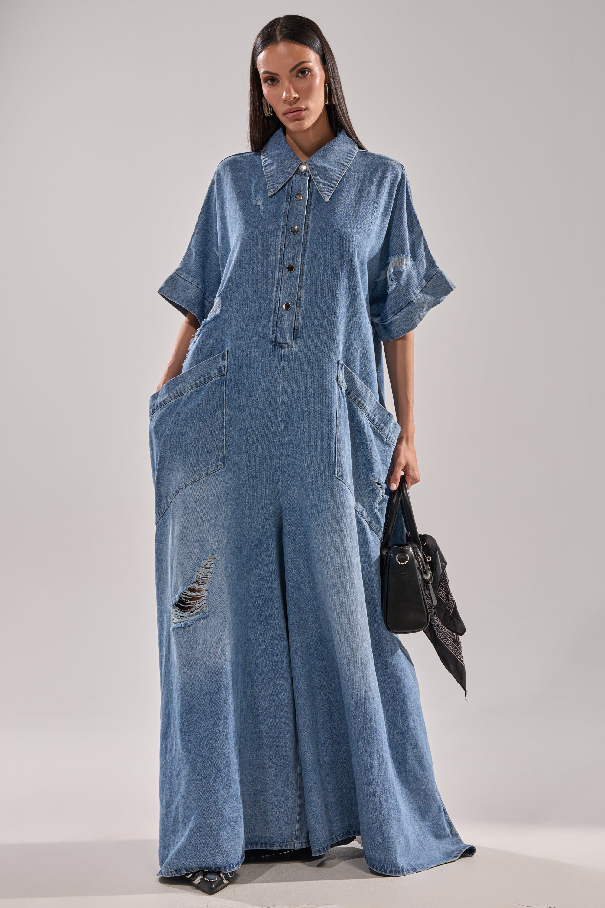 A woman wears THE BEST OVERSIZED DENIM JUMPSUIT with short sleeves, a large collar, button-down front, oversized pockets, and distressed details. She poses against a plain background while holding a black handbag.