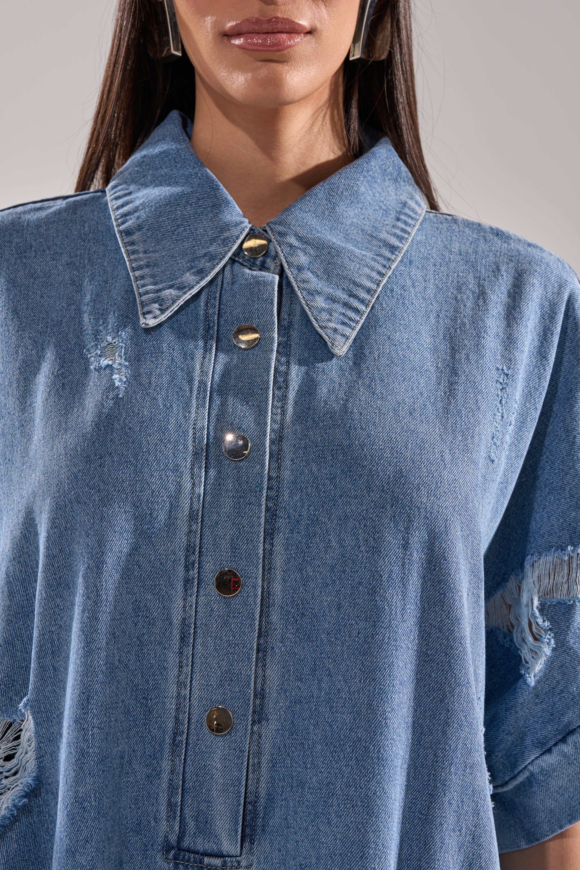 A close-up of a woman wearing THE BEST OVERSIZED DENIM JUMPSUIT in light blue, featuring a baggy fit, large collar, metal buttons, and distressed patches on the sleeves and chest. Only her lips and chin are visible.