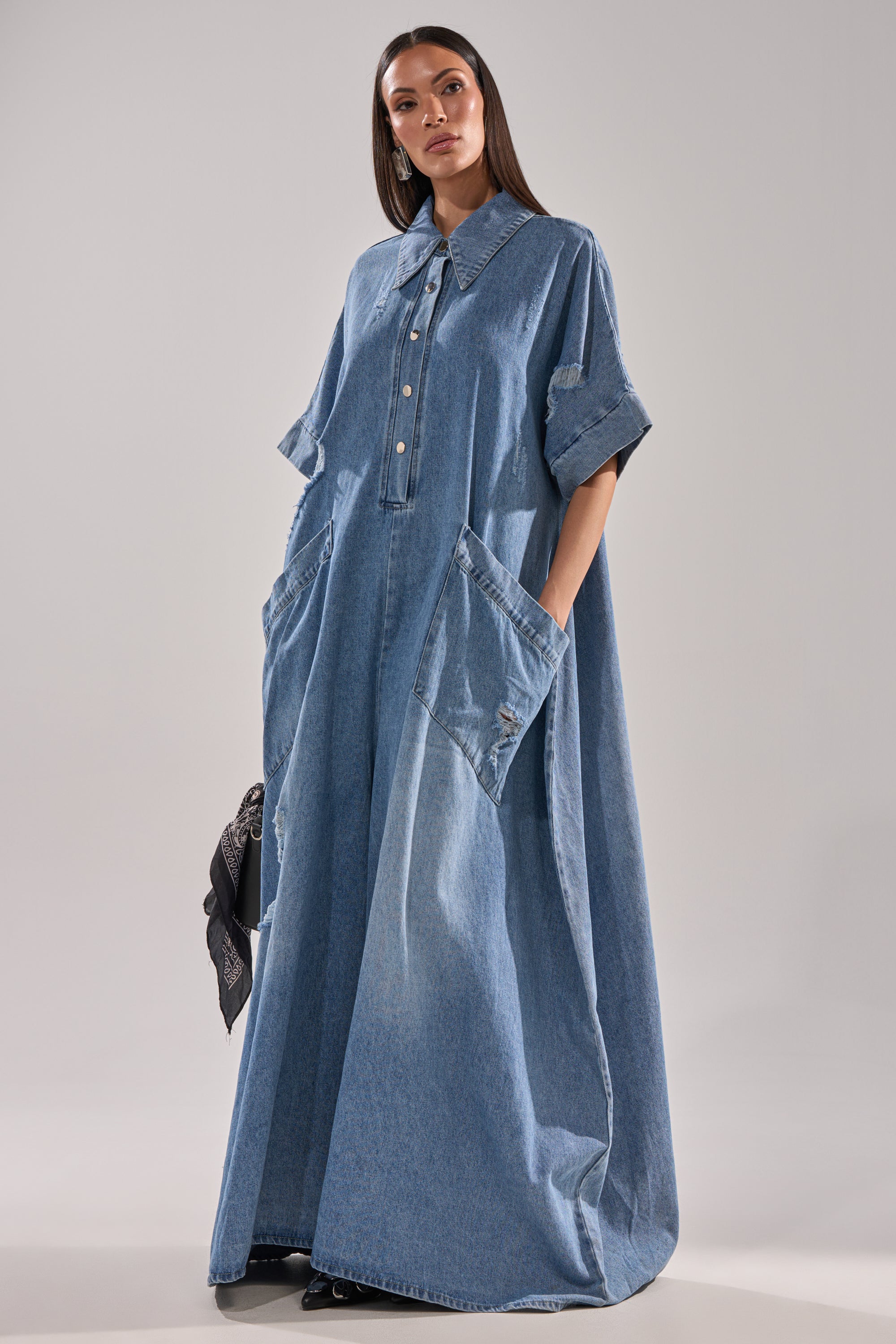 A woman models THE BEST OVERSIZED DENIM JUMPSUIT, a relaxed, floor-length piece with short sleeves, large front pockets, and a wide collar. She holds a black handbag and gazes confidently at the camera against a plain backdrop.