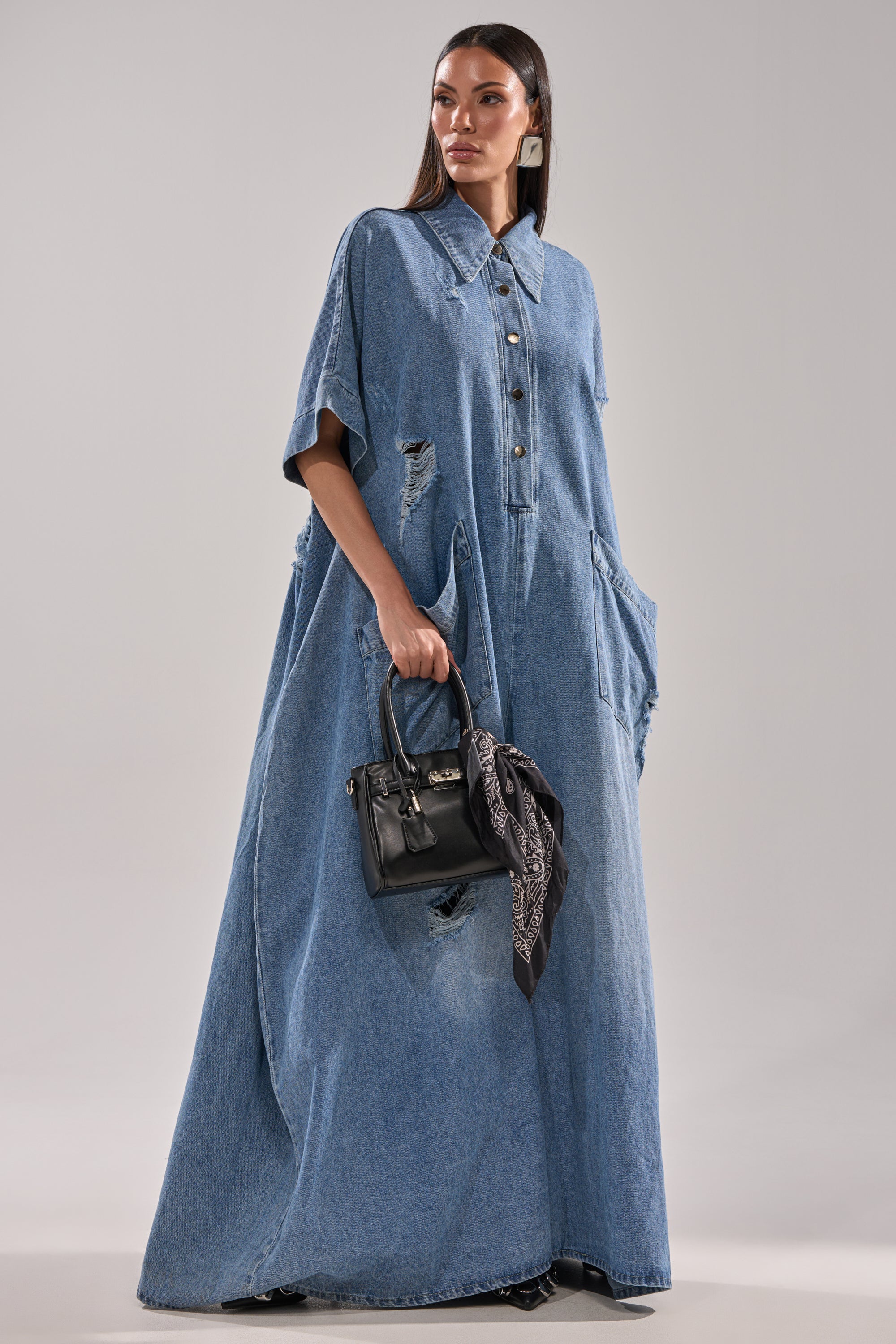 A woman models THE BEST OVERSIZED DENIM JUMPSUIT—a loose, floor-length short-sleeve piece with a relaxed baggy fit. She accessorizes with a small black handbag featuring a patterned scarf, standing before a plain gray background.