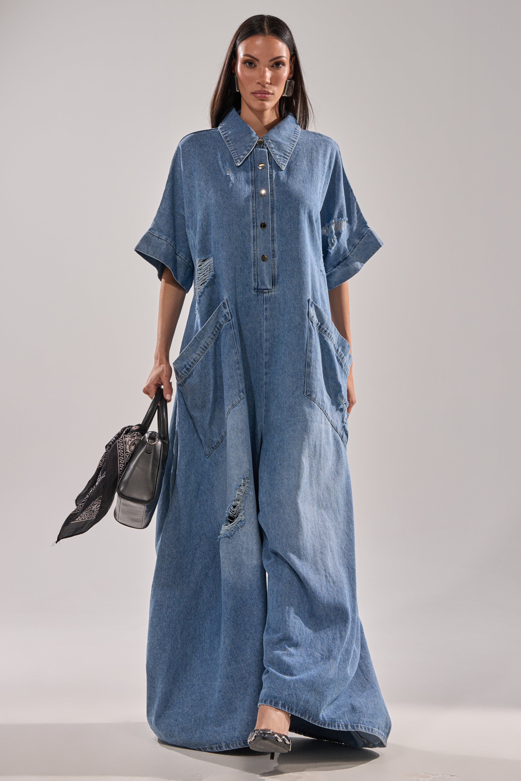 A woman with long, straight hair walks forward in THE BEST OVERSIZED DENIM JUMPSUIT—floor-length, relaxed fit, distressed denim with large front pockets and short sleeves. She holds a black handbag and wears heeled shoes.