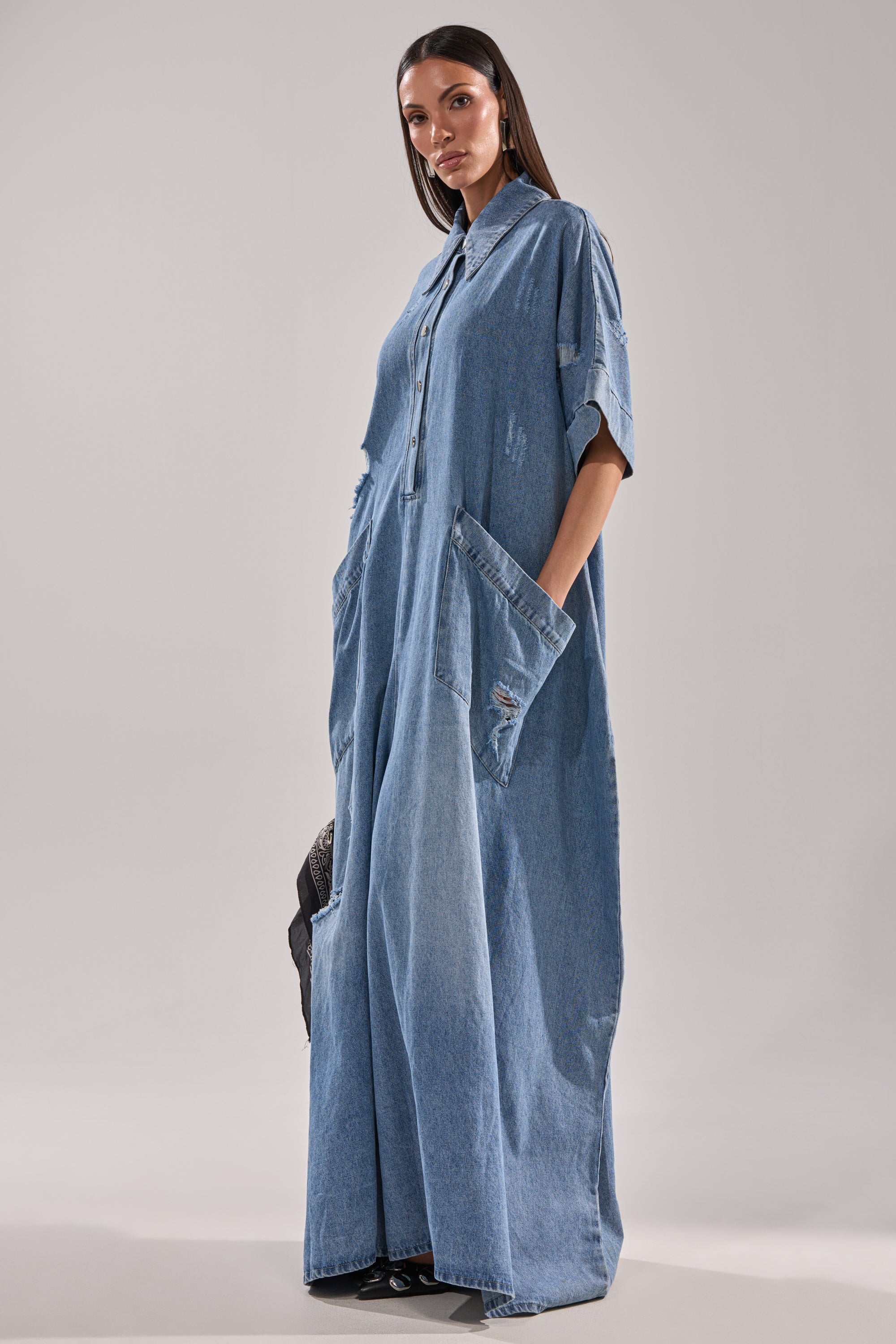 A woman wears THE BEST OVERSIZED DENIM JUMPSUIT, a loose-fitting, floor-length denim piece with short sleeves, collar, large front pockets, and a relaxed baggy fit, standing against a plain light background.