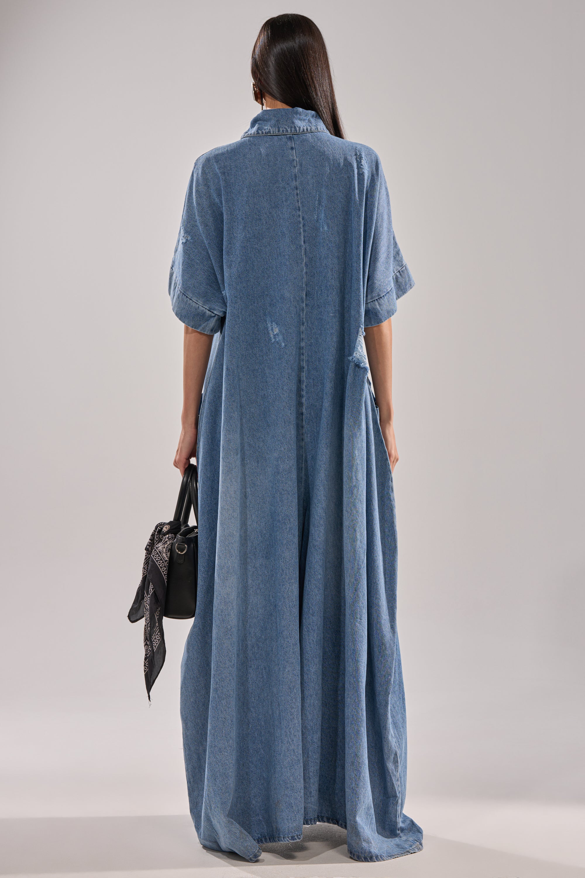 A person with long dark hair faces away in THE BEST OVERSIZED DENIM JUMPSUIT, a relaxed baggy light blue one-piece with sleeves. They hold a black handbag adorned with a black patterned scarf against a plain, neutral background.