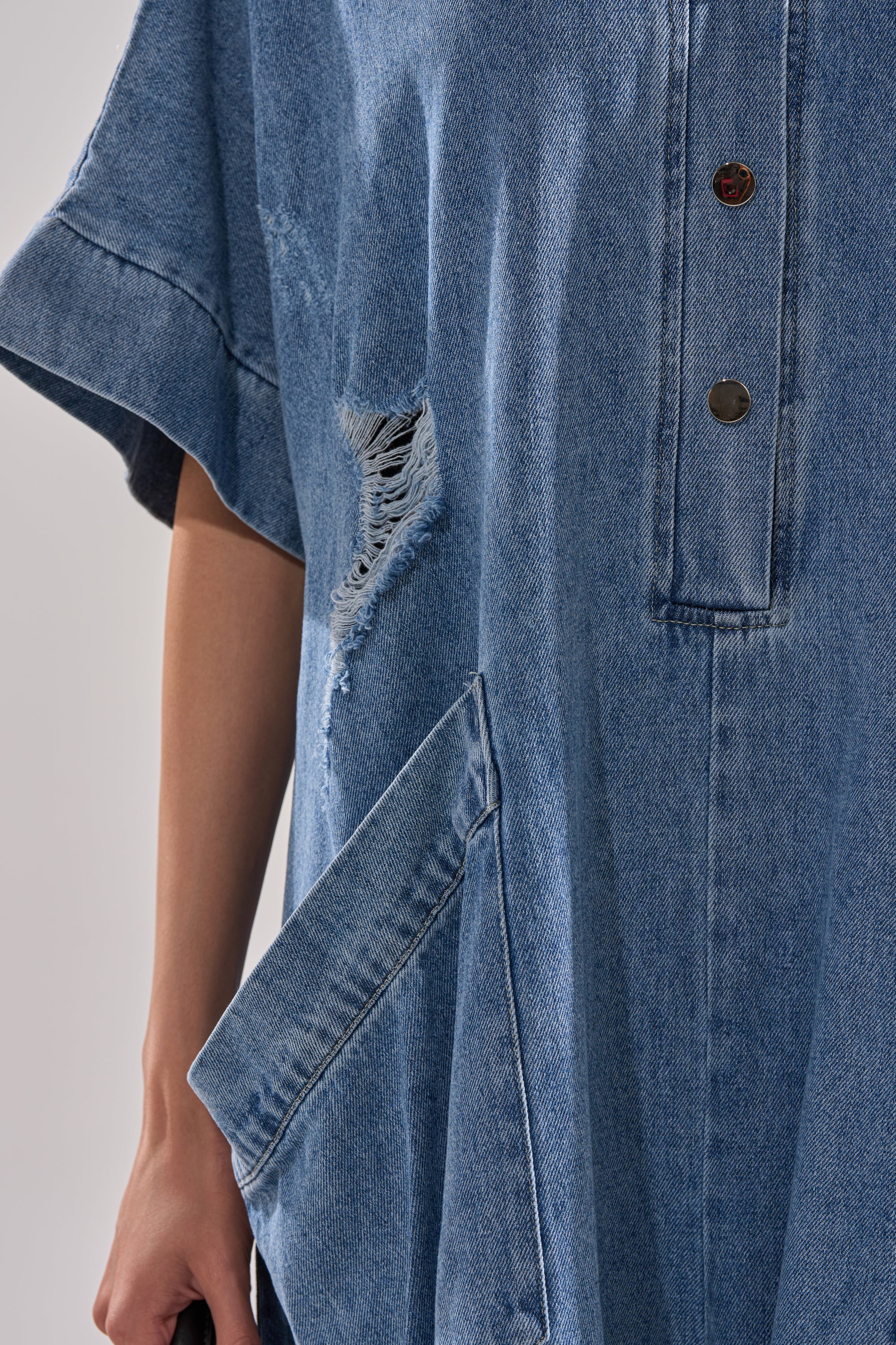 Model wears THE BEST OVERSIZED DENIM JUMPSUIT, featuring a ripped chest detail, metal snap buttons, a large front pocket, and classic distressed denim in a relaxed, baggy fit. Their left hand hangs casually by their side.