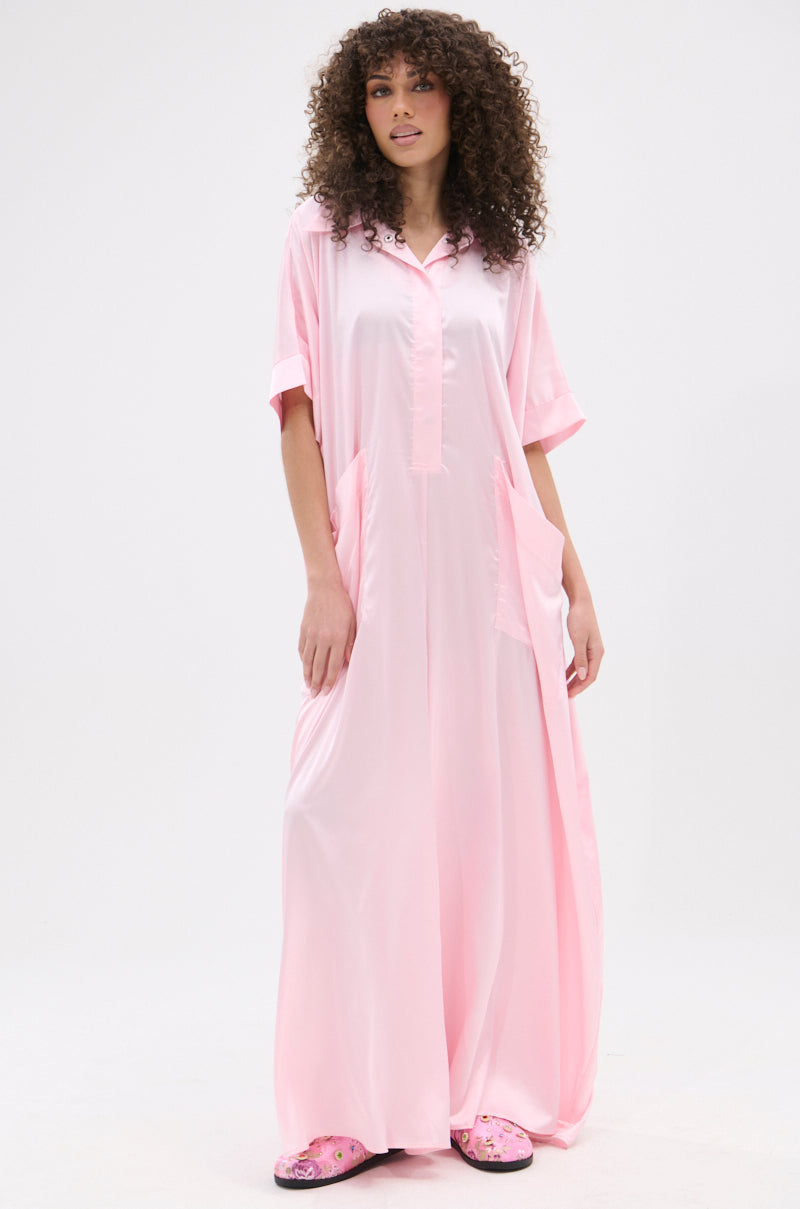 A woman with curly hair wears the SHE'S RICH OVERSIZED SATIN JUMPSUIT IN LIGHT PINK—luxurious loungewear with short sleeves and large pockets. She pairs it with floral pink slippers, standing against a plain white background.