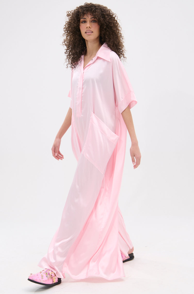 A woman with curly hair models the SHE'S RICH OVERSIZED SATIN JUMPSUIT IN LIGHT PINK, paired with pink floral slippers, against a plain white background.