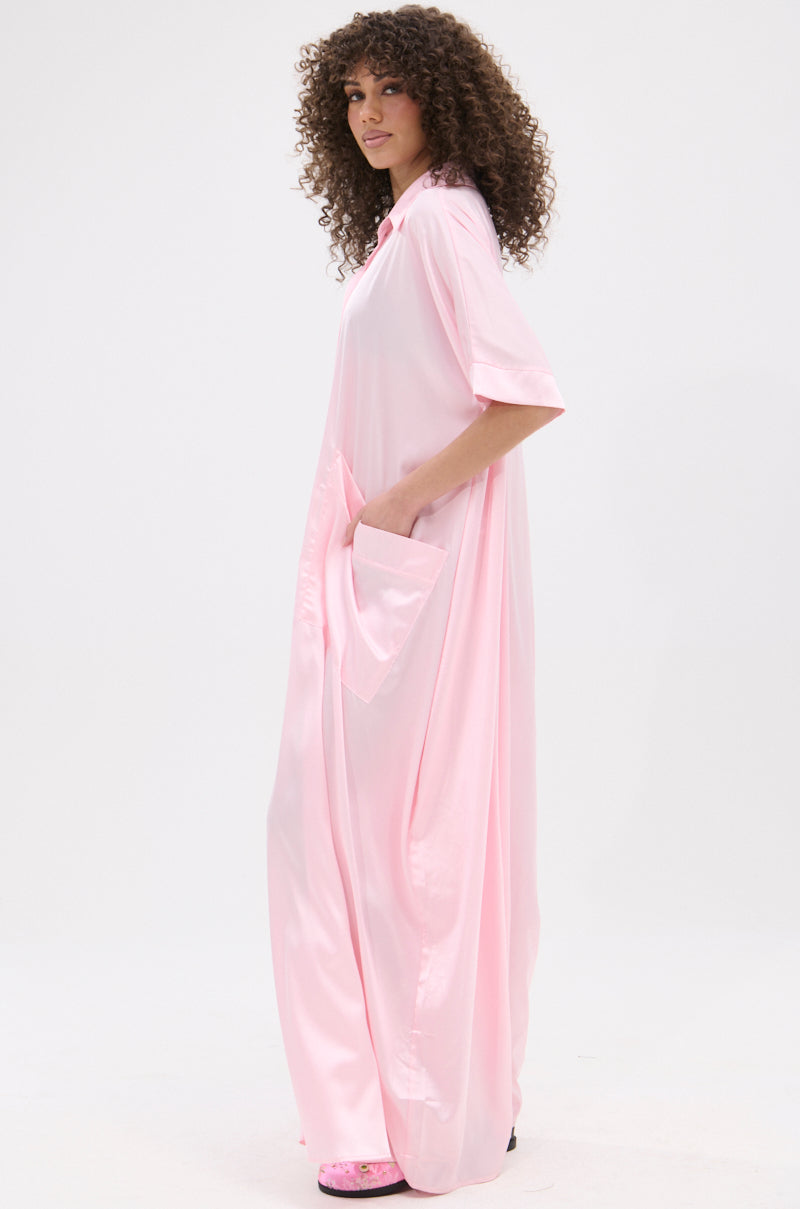 A person with curly hair stands sideways in the SHE'S RICH OVERSIZED SATIN JUMPSUIT IN LIGHT PINK, a loose, shiny one-piece with short sleeves and pockets. It reaches the floor and is styled with pink open-toe sandals against a plain white background.