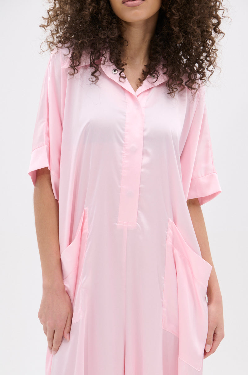 A woman with curly hair models the SHE'S RICH OVERSIZED SATIN JUMPSUIT IN LIGHT PINK, a loose short-sleeve one-piece with two large front pockets. The photo, cropped at her shoulders and knees, shows effortless vacation style against a plain background.