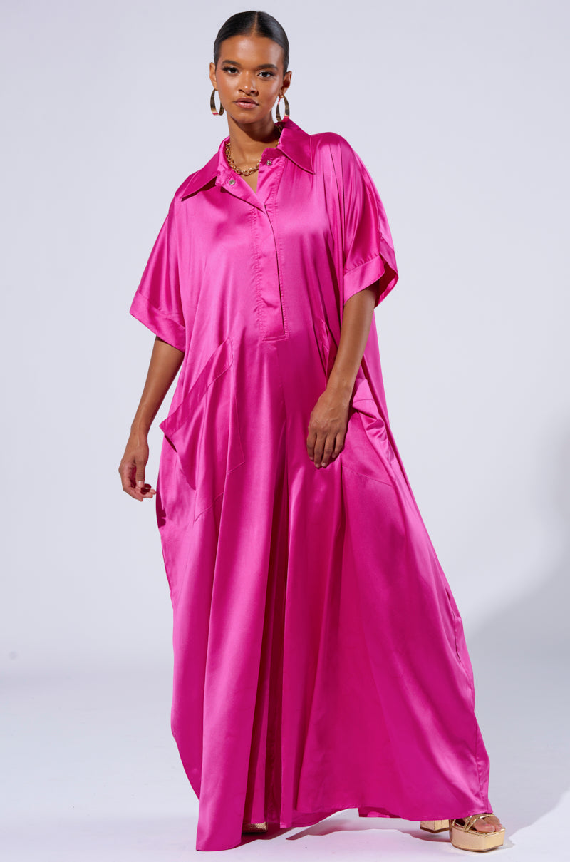 A woman confidently models the SHES RICH OVERSIZED SATIN JUMPSUIT IN PINK, featuring a vibrant, hot pink color and wide legs with short sleeves. She accessorizes with large hoop earrings and a necklace. Her hair is pulled back as she stands against a plain white background.