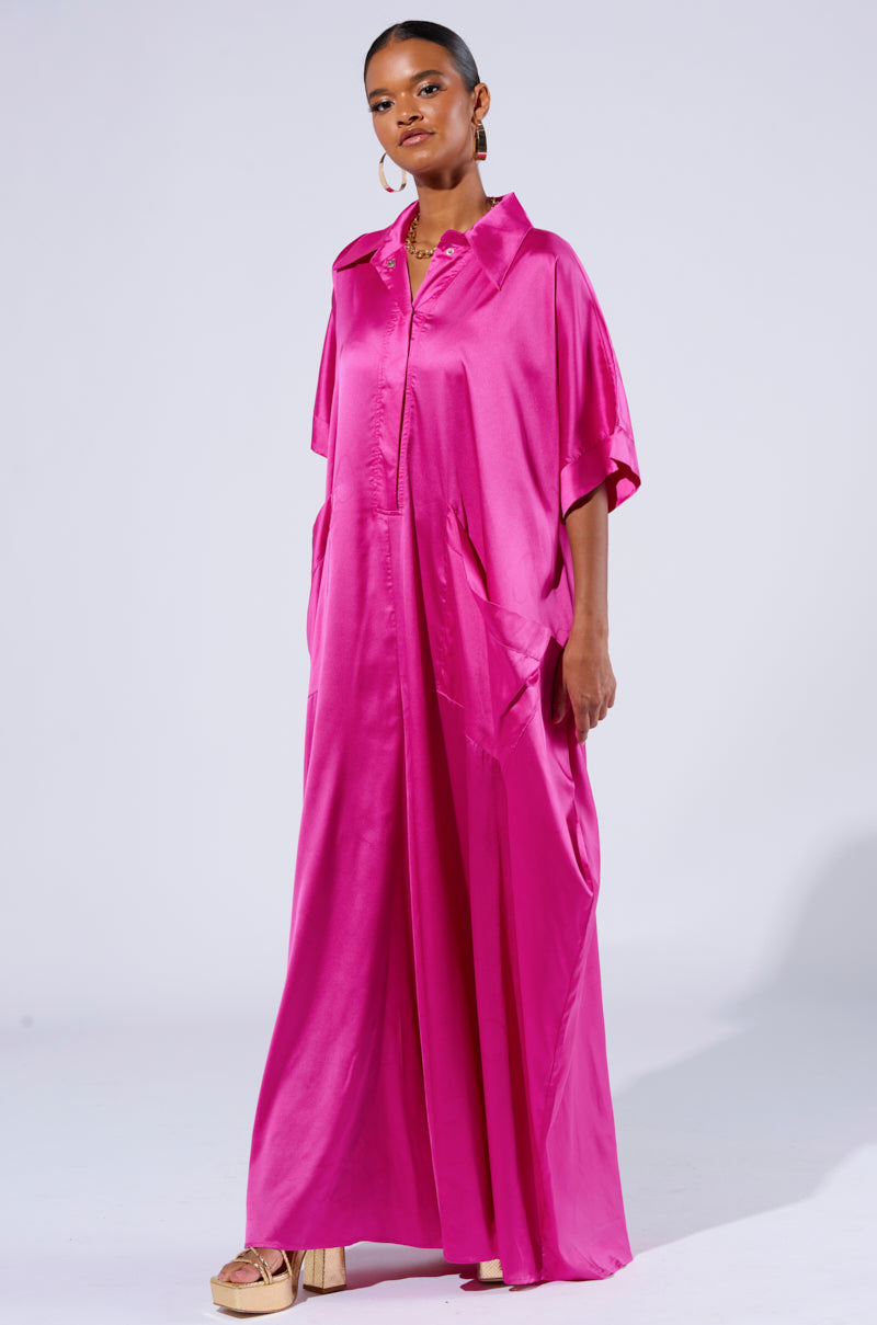 A woman stands confidently in the SHES RICH OVERSIZED SATIN JUMPSUIT IN PINK, featuring short sleeves and a flowing silhouette. She has her hands in the jumpsuit's pockets and is wearing beige platform sandals. She has hoop earrings and her hair is neatly pulled back. The background is plain white.