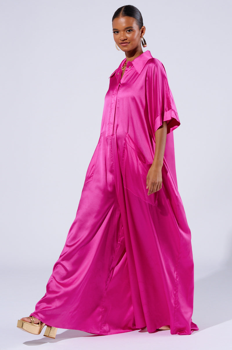 A person models the SHES RICH OVERSIZED SATIN JUMPSUIT IN PINK, featuring short sleeves, a collar, and button-down details. The wide-leg jumpsuit boasts a flowing silhouette and two large pockets. They complete the look with gold platform sandals and large hoop earrings against a neutral background.