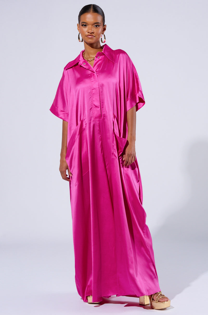 A woman is standing and wearing the SHES RICH OVERSIZED SATIN JUMPSUIT IN PINK. She accessorizes with hoop earrings, a necklace, and a pair of sandals. The background is plain and light-colored.