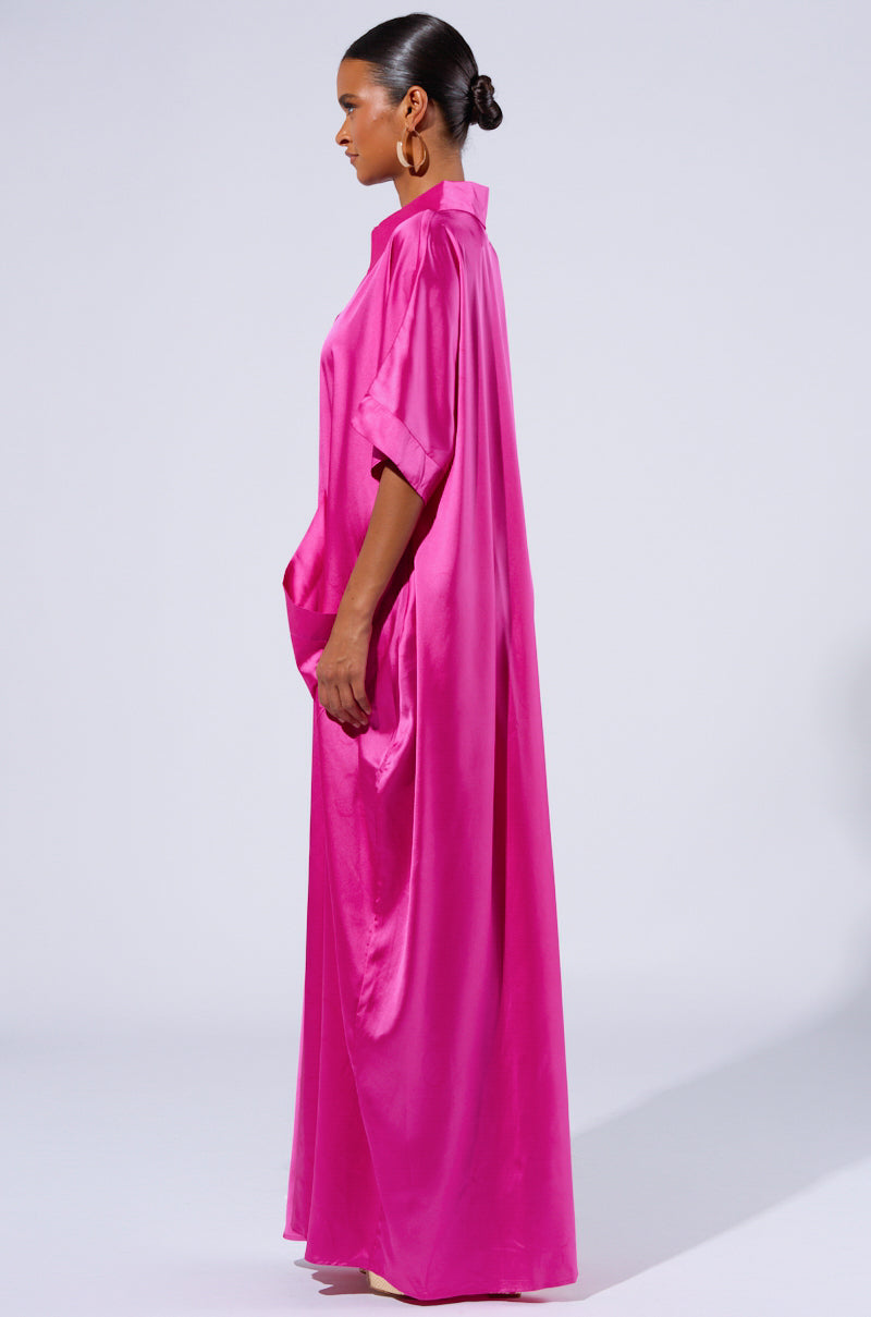 A woman stands in profile, wearing the SHES RICH OVERSIZED SATIN JUMPSUIT IN PINK. The jumpsuit features a collar, short sleeves, and wide legs that reach the floor. She has her hair tied back in a neat bun and wears large hoop earrings. The background is plain and light-colored.