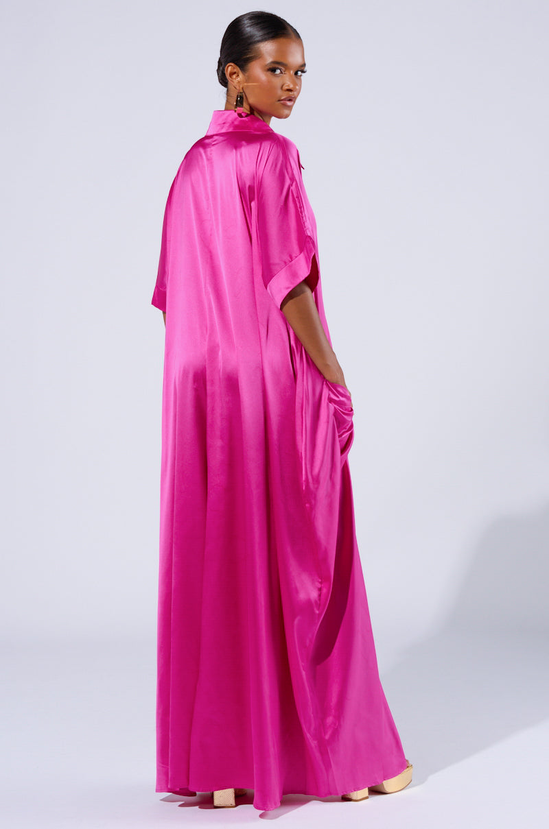 A person stands facing slightly towards the back left, wearing the "SHES RICH OVERSIZED SATIN JUMPSUIT IN PINK." The jumpsuit shines in the light, highlighting its smooth, silky texture similar to a vibrant magenta gown. The person has short hair, and the background is a neutral gray.