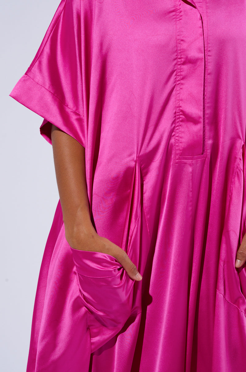 A person is wearing the SHES RICH Oversized Satin Jumpsuit in Pink, which boasts a vibrant hue and an oversized fit. The jumpsuit features short sleeves and pockets, crafted from smooth fabric with a noticeable sheen. With their right hand partially tucked into one of the pockets, the relaxed and chic design is accentuated beautifully.
