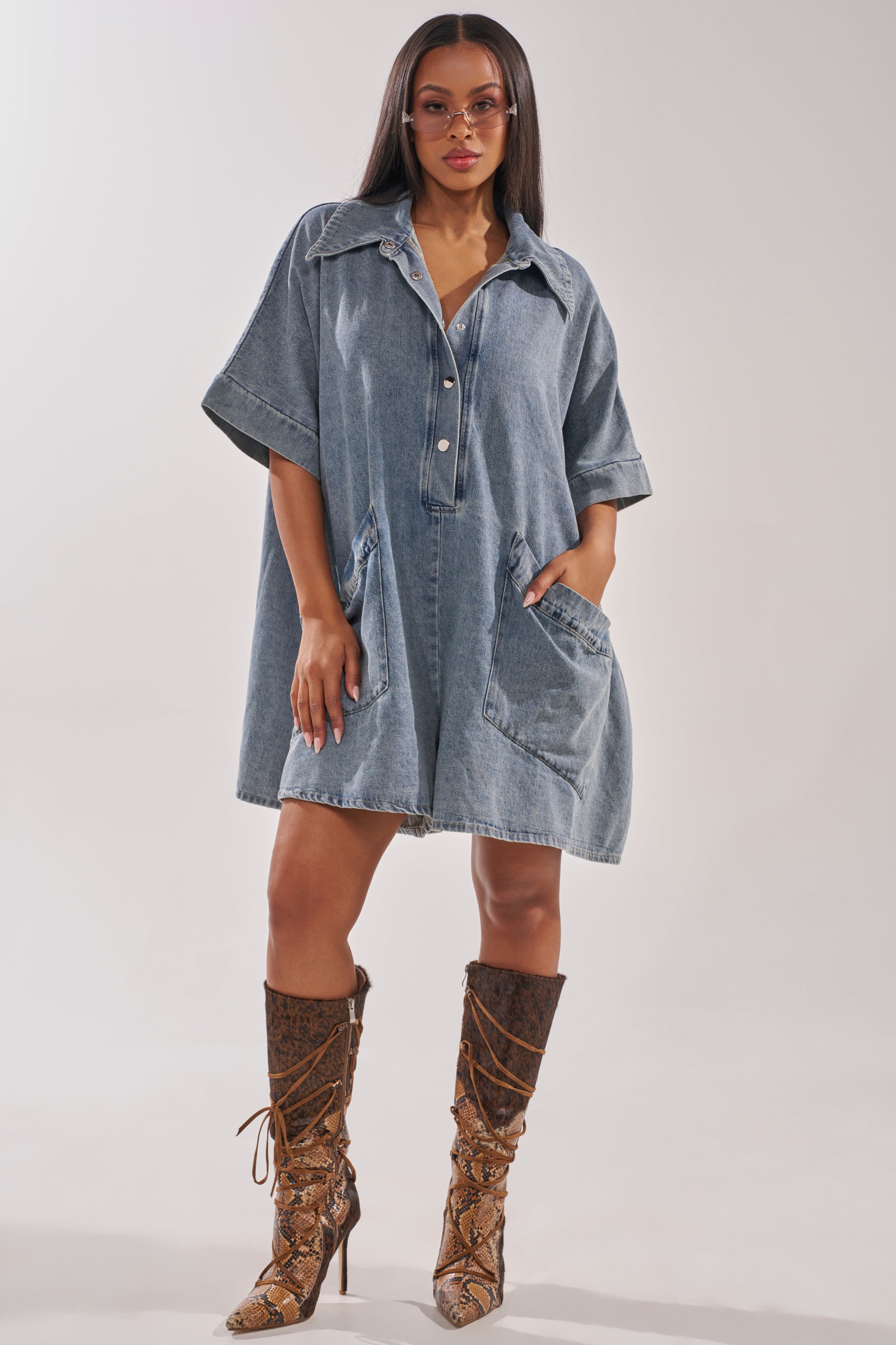 A woman wears the MELODY COLLARED DENIM ROMPER with short sleeves and large pockets, styled with lace-up snakeskin knee-high boots and pink tinted sunglasses. She stands confidently against a plain backdrop. 0