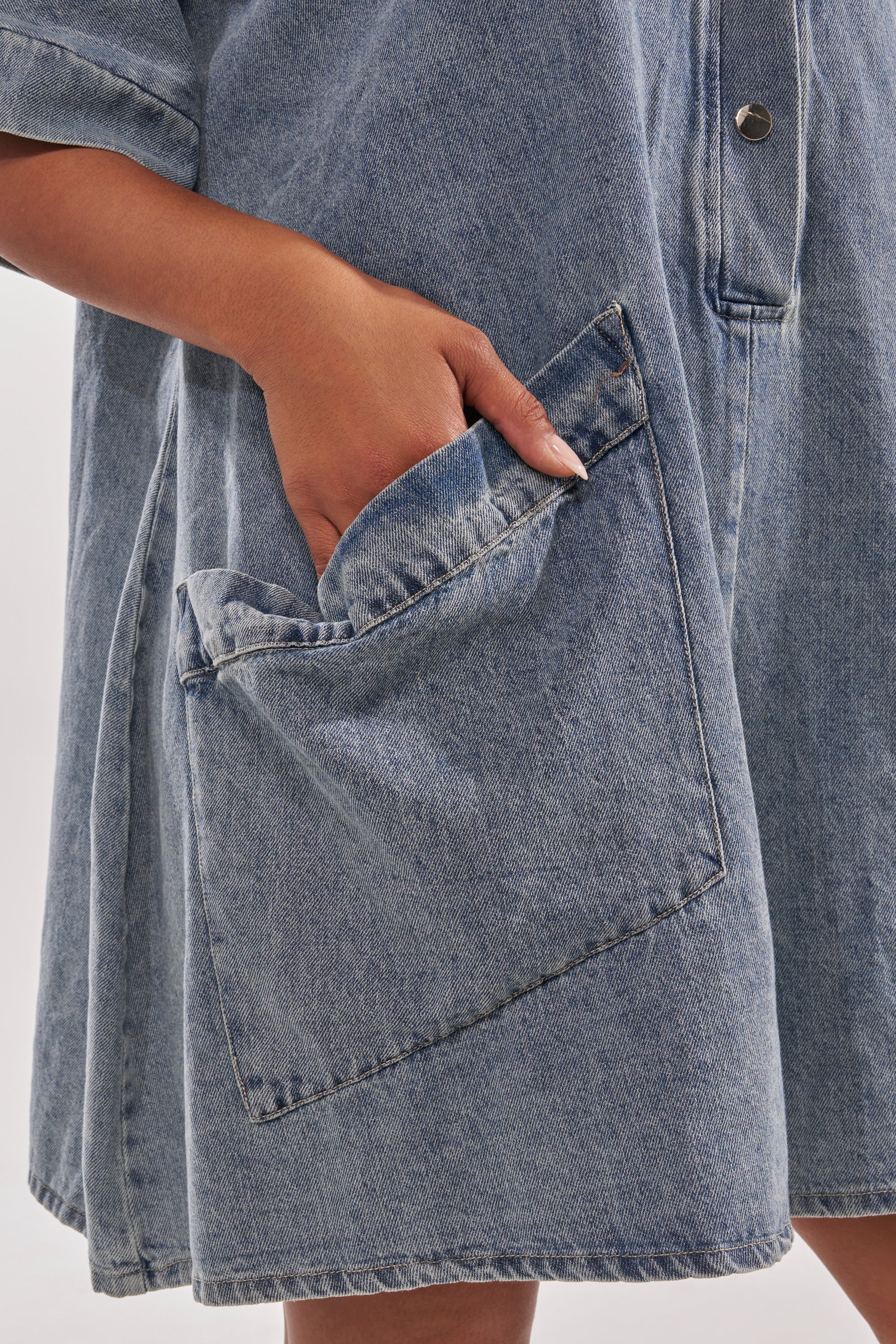 A person wears the MELODY COLLARED DENIM ROMPER, a blue street style outfit featuring a collar and large angled front pockets, with their hand partially tucked into one pocket. 9