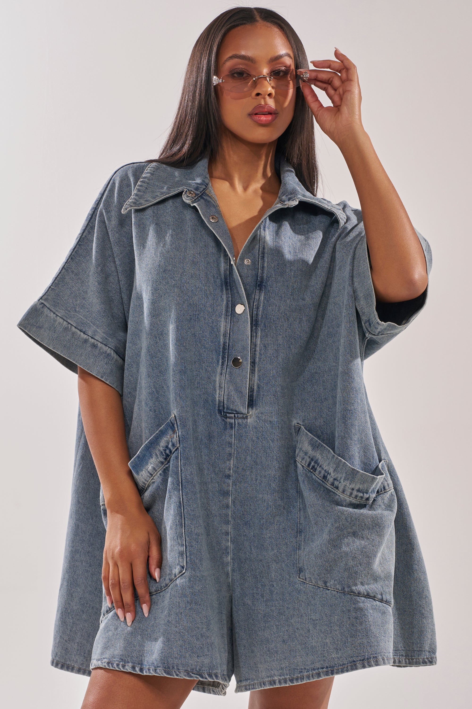 A woman poses confidently against a plain background, wearing the MELODY COLLARED DENIM ROMPER—an oversized denim romper with front buttons and two large front pockets—while touching her sunglasses with one hand. 1