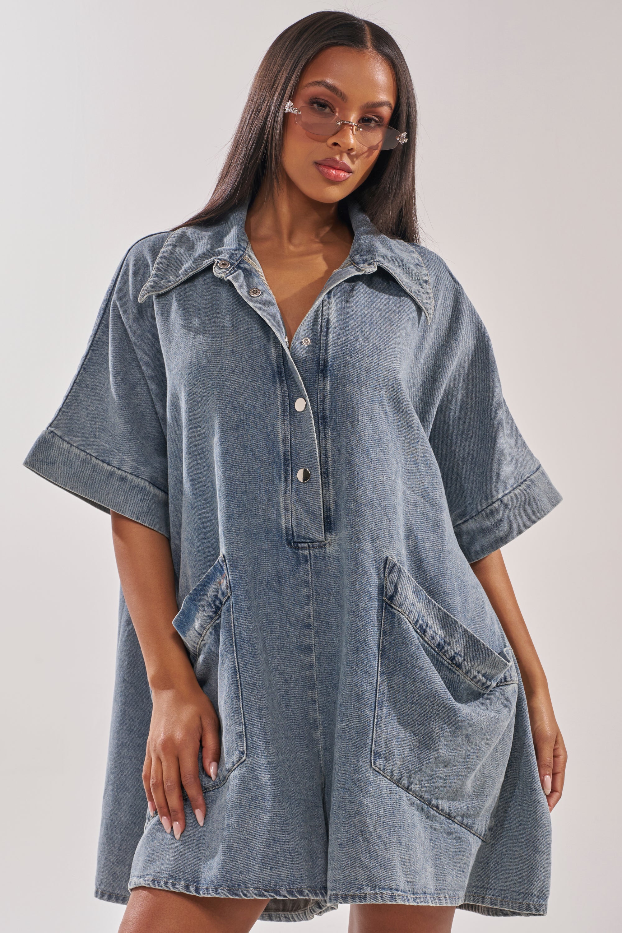 A woman models the MELODY COLLARED DENIM ROMPER, featuring short sleeves and large front pockets. Paired with light pink sunglasses, she stands against a simple background with one hand in her pocket for a casual vibe. 2