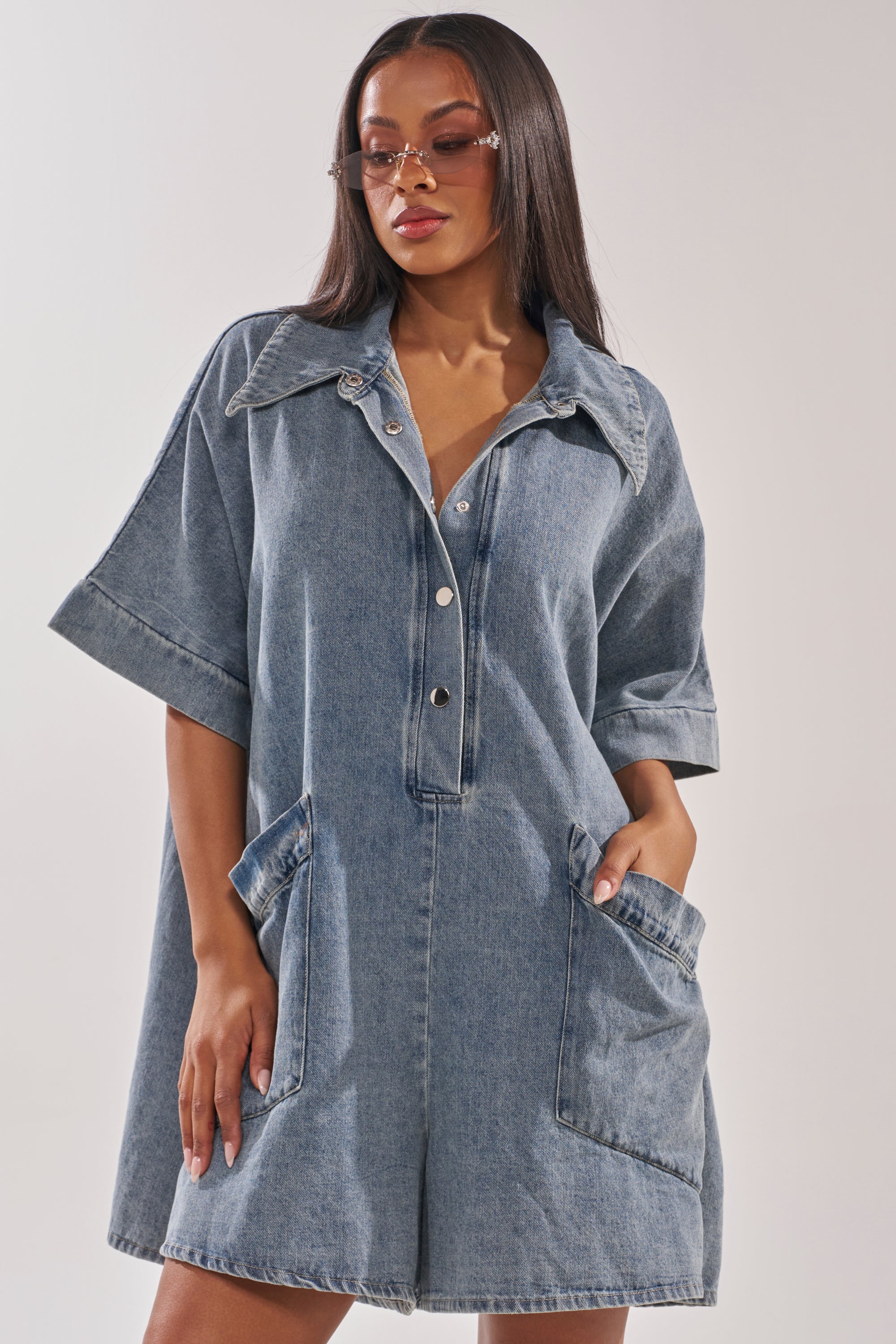 A woman models the MELODY COLLARED DENIM ROMPER with large front pockets and oversized sunglasses, hands tucked in, against a plain background for a relaxed street-style vibe. 3