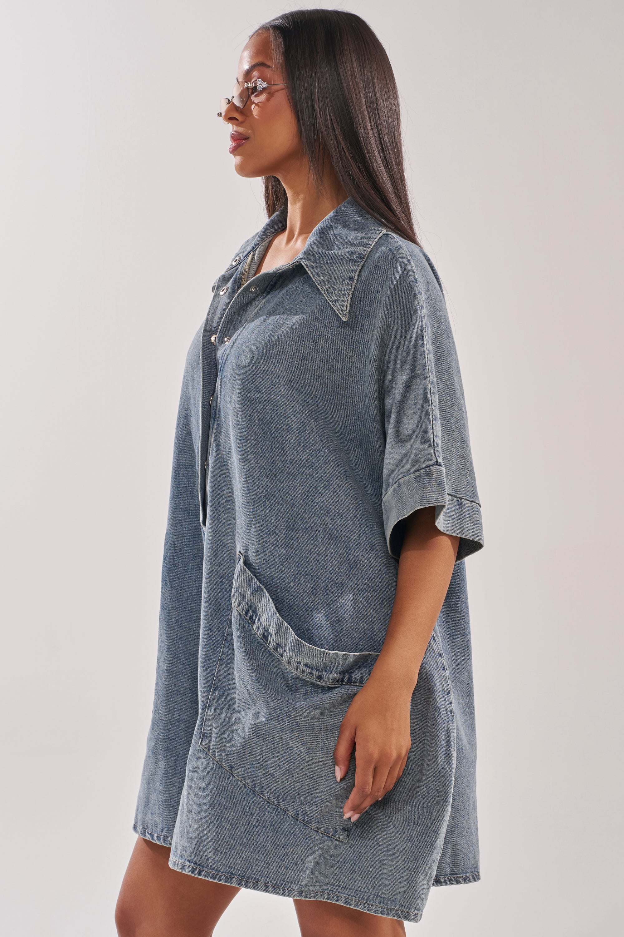 A woman with long, straight hair and glasses stands in profile against a plain light background, showcasing the oversized MELODY COLLARED DENIM ROMPER with its standout large collar and roomy pockets for an effortless street-style look. 4