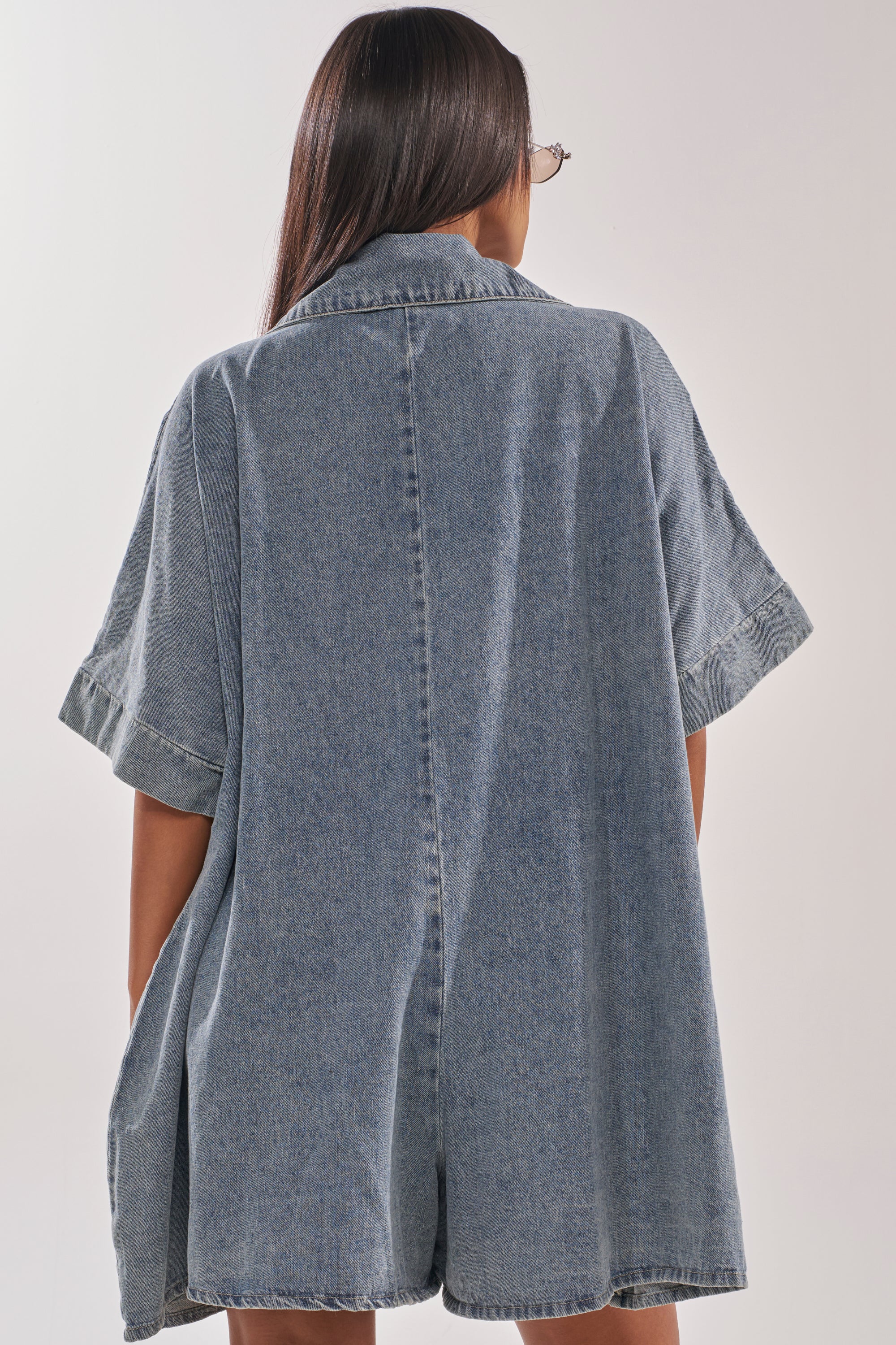 A person with long dark hair, wearing sunglasses and the MELODY COLLARED DENIM ROMPER, is seen from behind against a light background. 5