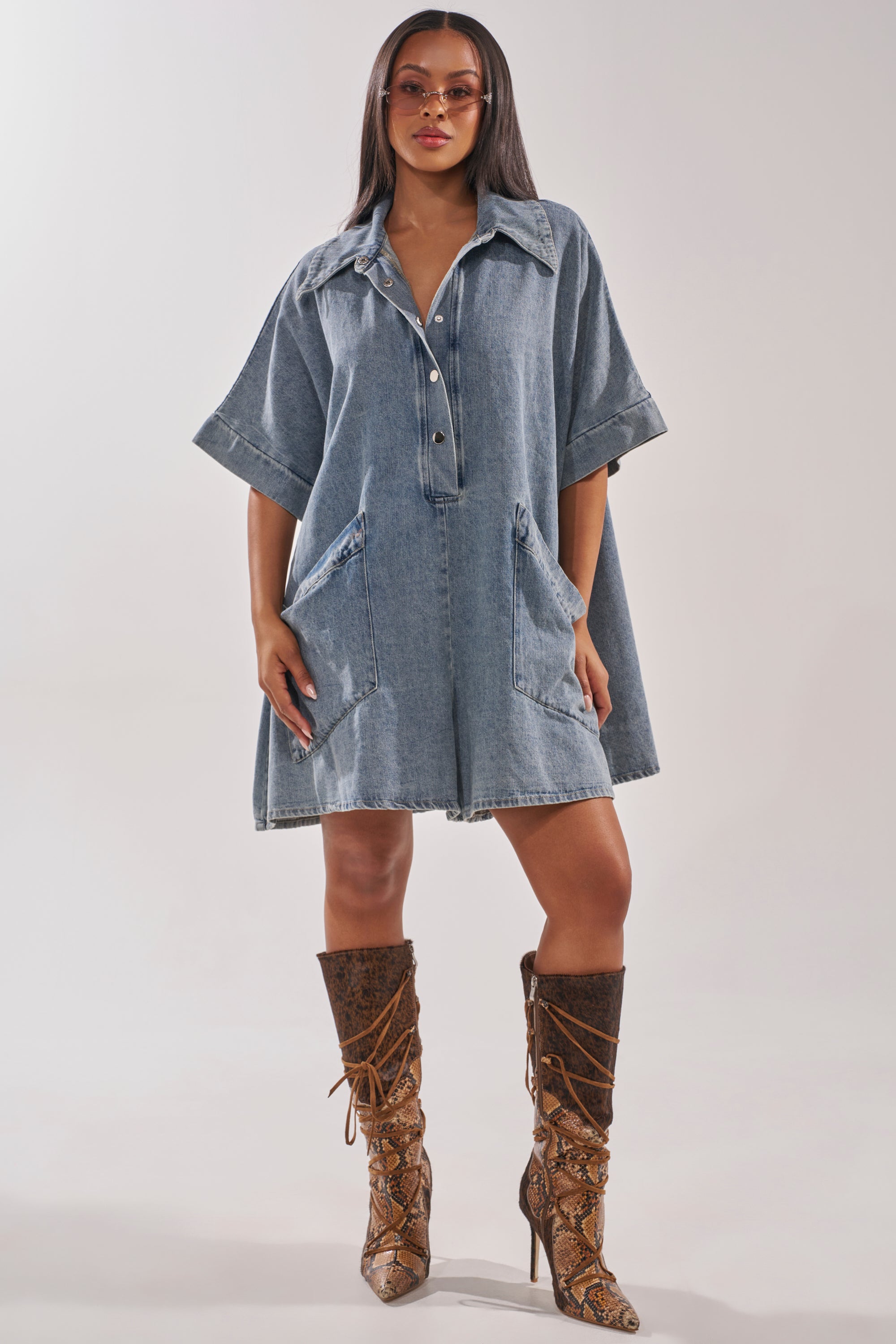 A woman exudes confidence in the MELODY COLLARED DENIM ROMPER with large pockets and snap buttons, styled with knee-high brown snakeskin lace-up boots and sunglasses for a chic, effortless street look. 6