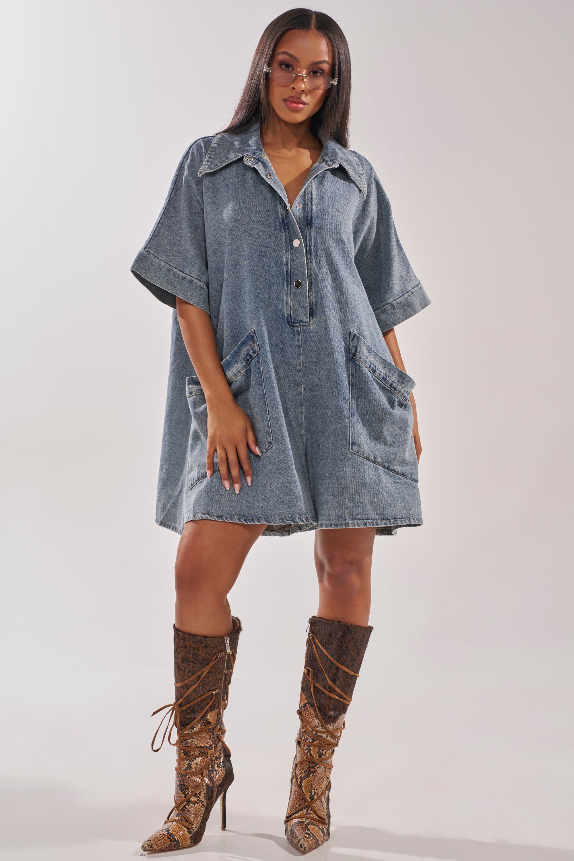 A woman exudes confidence in the MELODY COLLARED DENIM ROMPER with large front pockets, styled with knee-high lace-up snakeskin boots and clear-frame glasses for an effortlessly chic street style look. 7