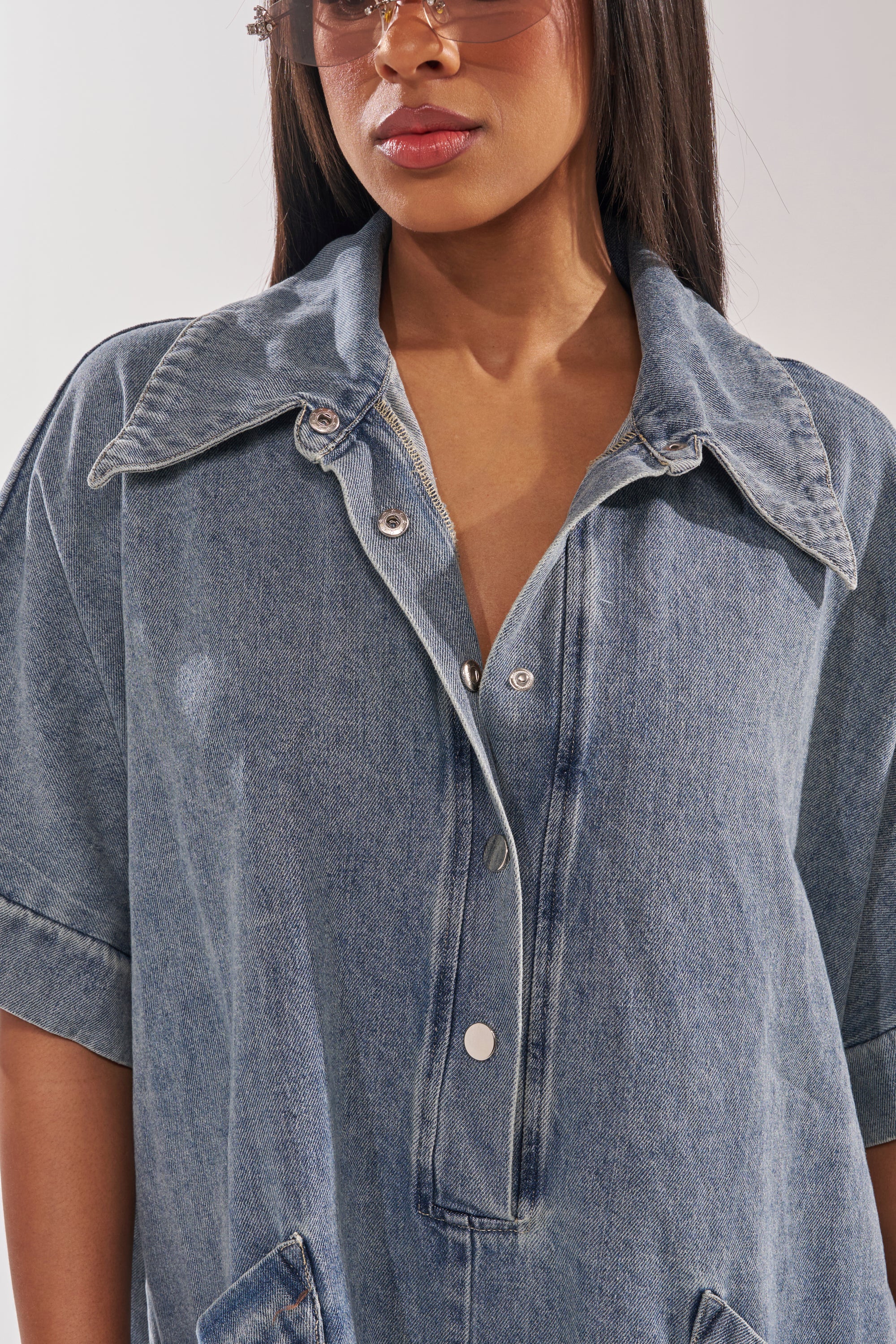 A woman wears the MELODY COLLARED DENIM ROMPER—an oversized style with a large collar and rolled sleeves—paired with light brown sunglasses. The cropped photo spotlights her effortless street style. 8