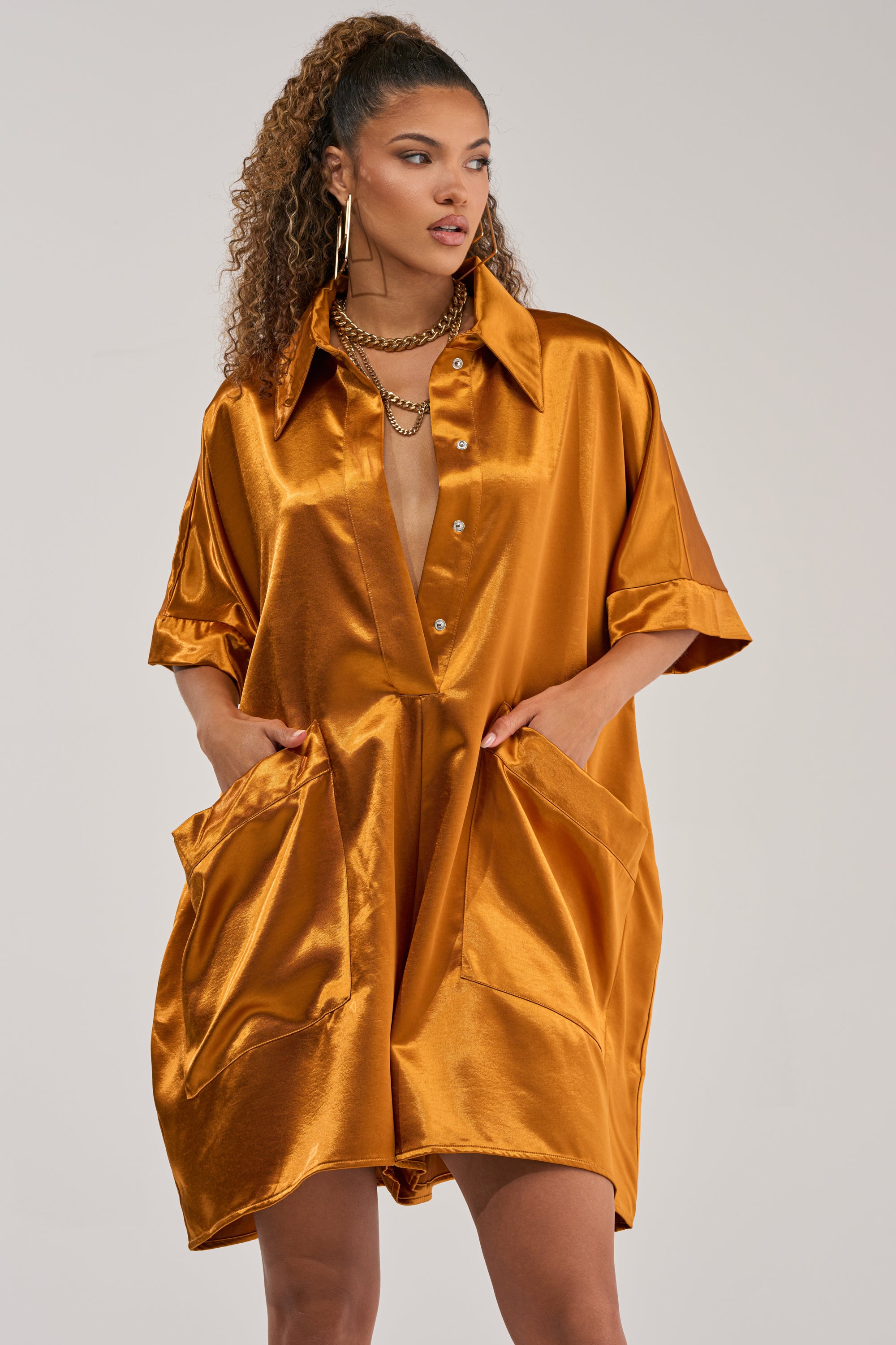 Wearing the SHE'S RICH OVERSIZED SATIN ROMPER IN MUSTARD—an effortlessly chic, loose-fitting piece featuring a lustrous finish, large front pockets, and rolled sleeves—a woman poses with her hands in her pockets against a plain background, embodying bold statement occasion style.