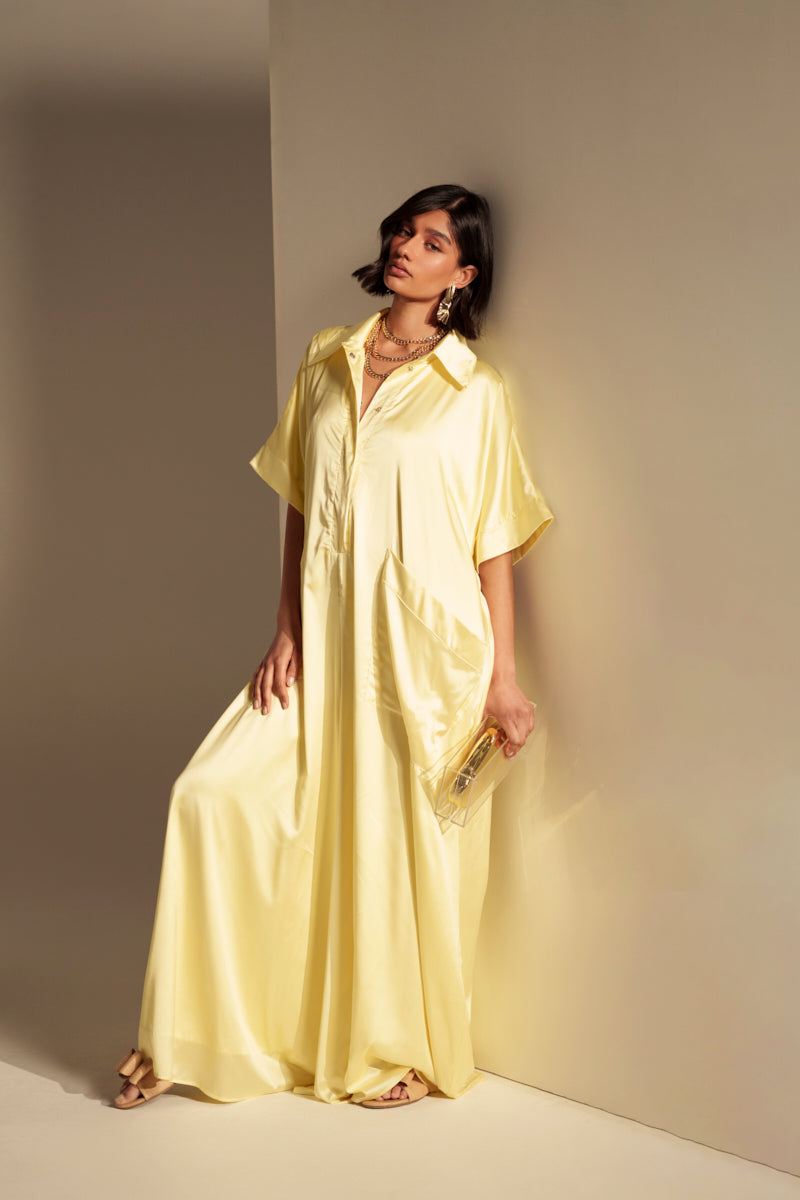 A woman in the HAPPY HOUR SATIN JUMPSUIT IN YELLOW, featuring a flowing wide-leg silhouette and short sleeves, leans against a beige wall with a clutch. She has a bob haircut, wears sandals and layered necklaces, and gazes to the side.