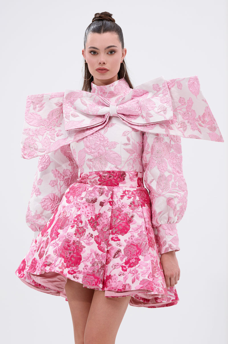 A woman wears the RENAISSANCE TWO-TONE BROCADE MINI DRESS IN PINK, featuring voluminous sleeves, a sculptural chest bow, and a pleated floral jacquard skirt, standing against a plain white background.