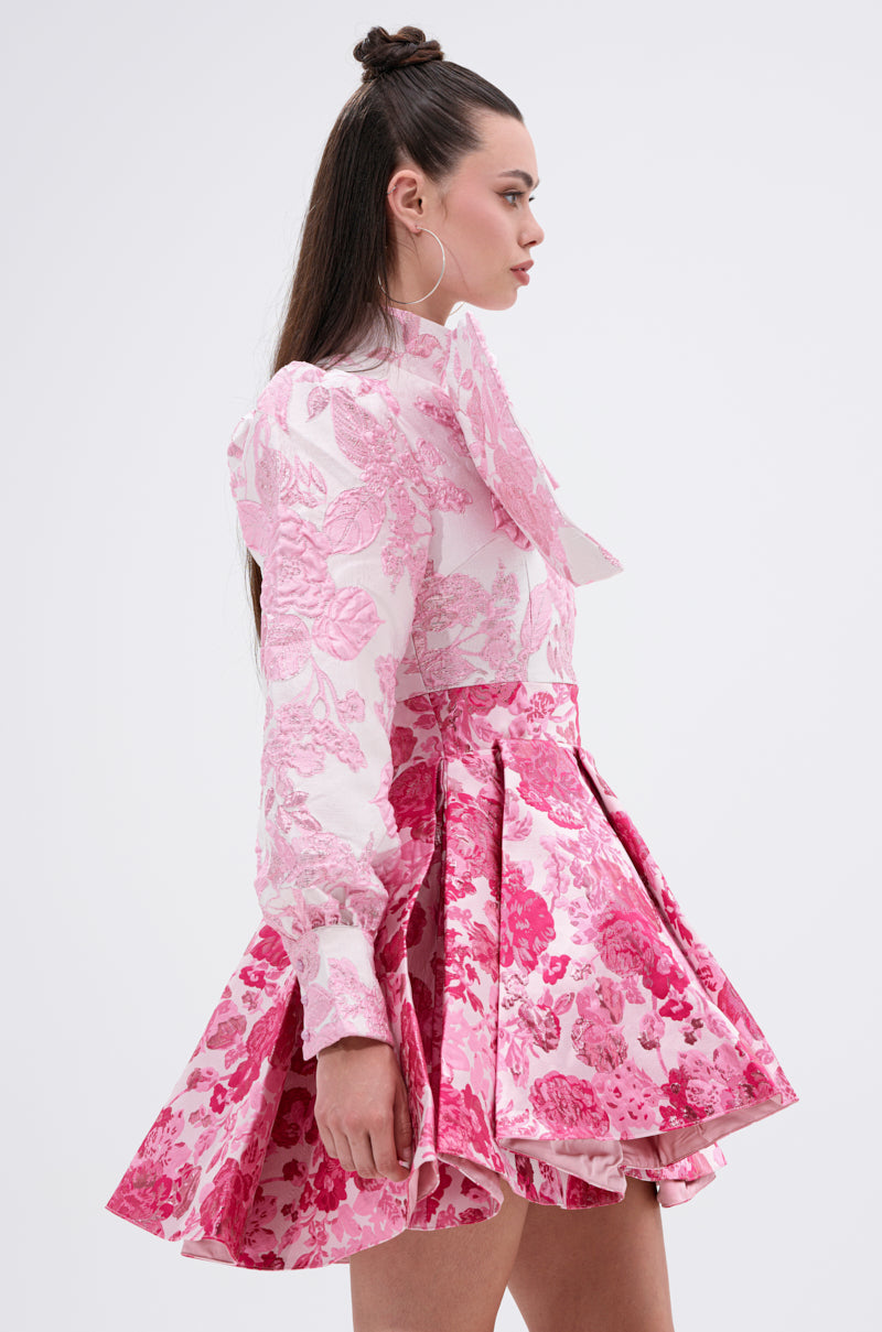 Woman in the RENAISSANCE TWO-TONE BROCADE MINI DRESS IN PINK, featuring pink and white floral jacquards, long sleeves, a fitted bodice, and flared skirt. She wears large hoop earrings; her long straight hair is partially pulled up.
