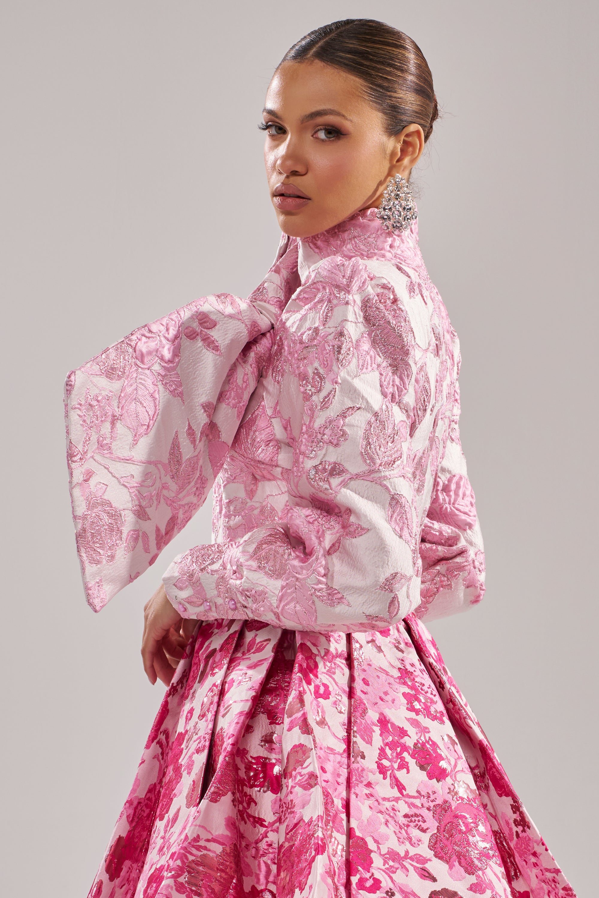 A woman poses against a plain background in the RENAISSANCE TWO-TONE BROCADE MINI DRESS IN PINK, featuring dramatic puff sleeves. She looks over her shoulder, wears statement earrings, and has her hair slicked back. 2