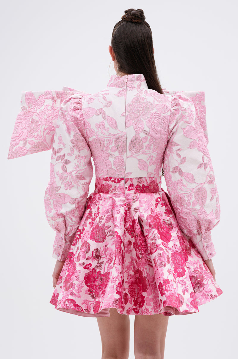 A woman faces away, wearing the RENAISSANCE TWO-TONE BROCADE MINI DRESS IN PINK with puffy sleeves, sculptural shoulder bows, a fitted bodice, and a flared skirt featuring bold floral jacquard patterns.