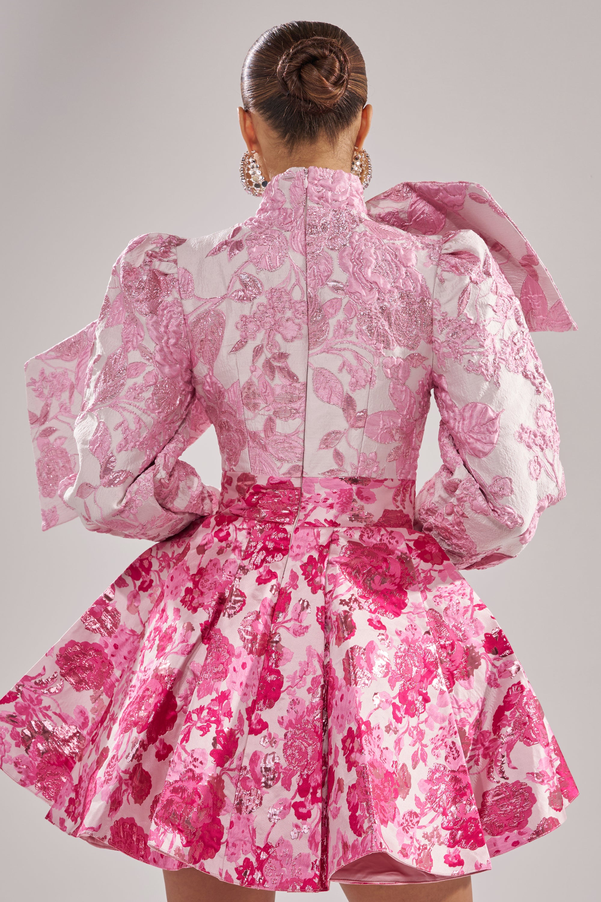 A person with hair in a neat bun wears the RENAISSANCE TWO-TONE BROCADE MINI DRESS IN PINK, featuring long sleeves, a large shoulder bow, and flared skirt. Seen from the back as they face away from the camera. 3
