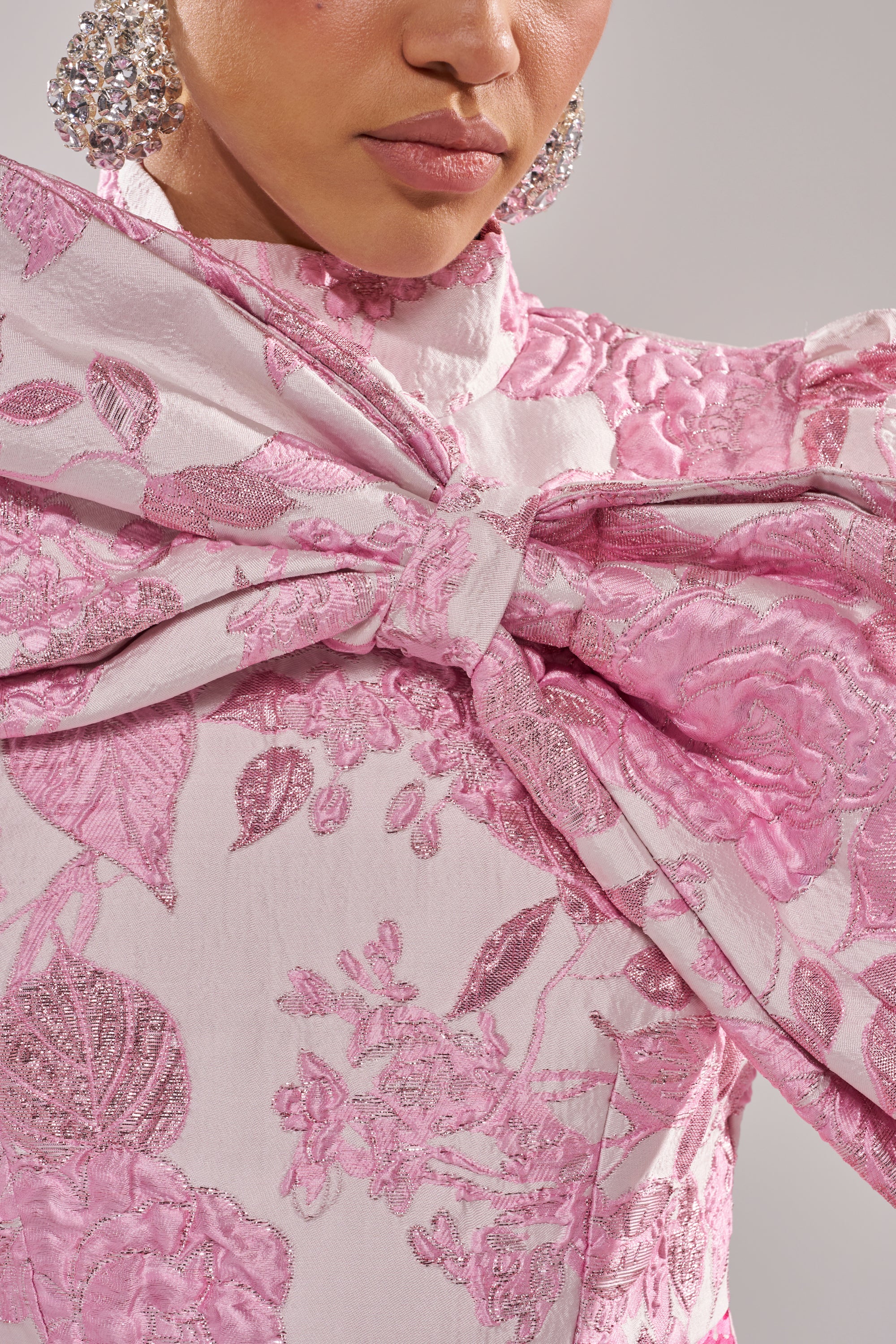 A close-up of a person wearing the RENAISSANCE TWO-TONE BROCADE MINI DRESS IN PINK, featuring textured patterns and a large chest bow. Rhinestone earrings and pink lipstick complete the elegant look. 4